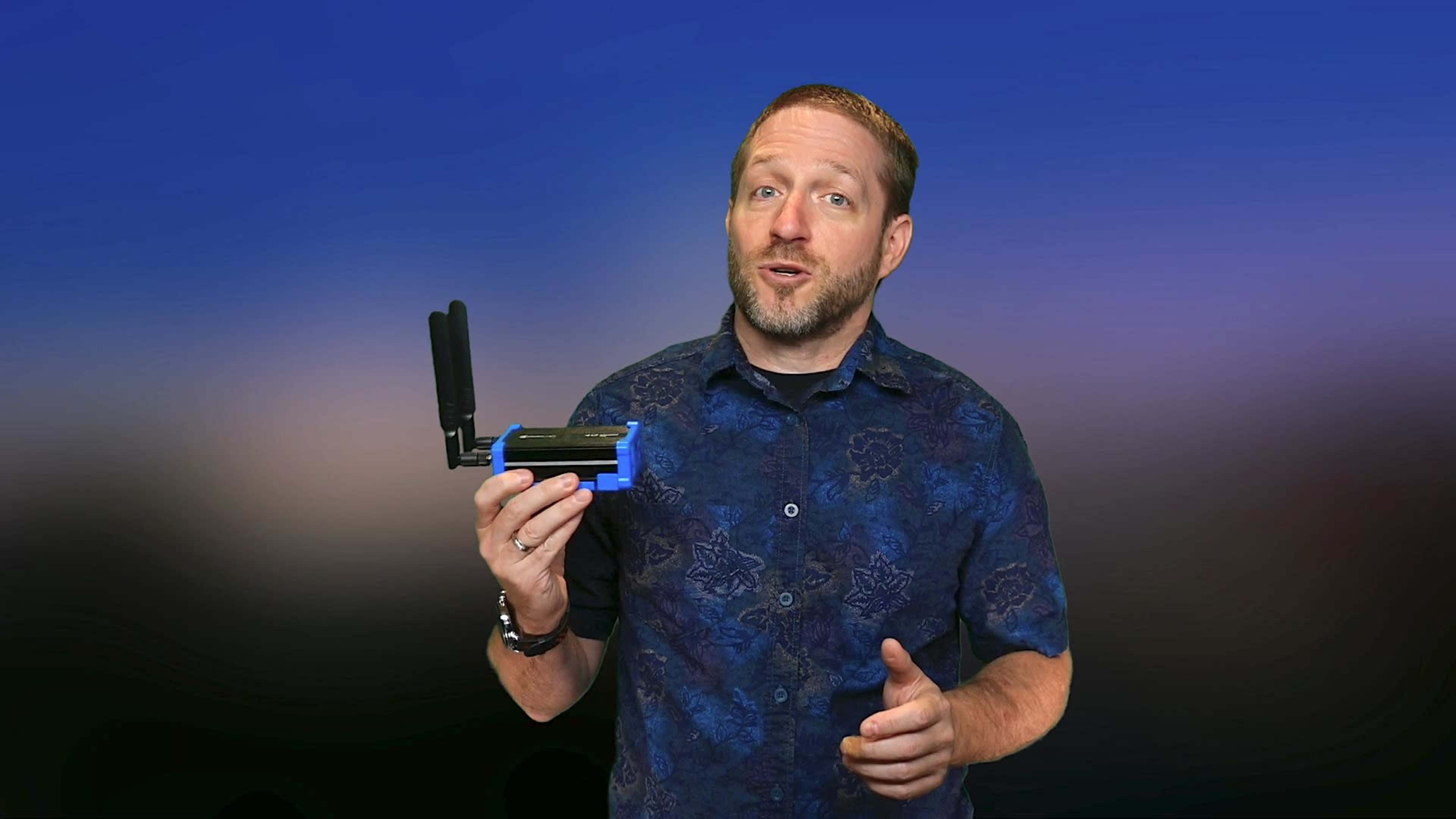 A man holds a small electronic device with antennas while speaking towards the camera.