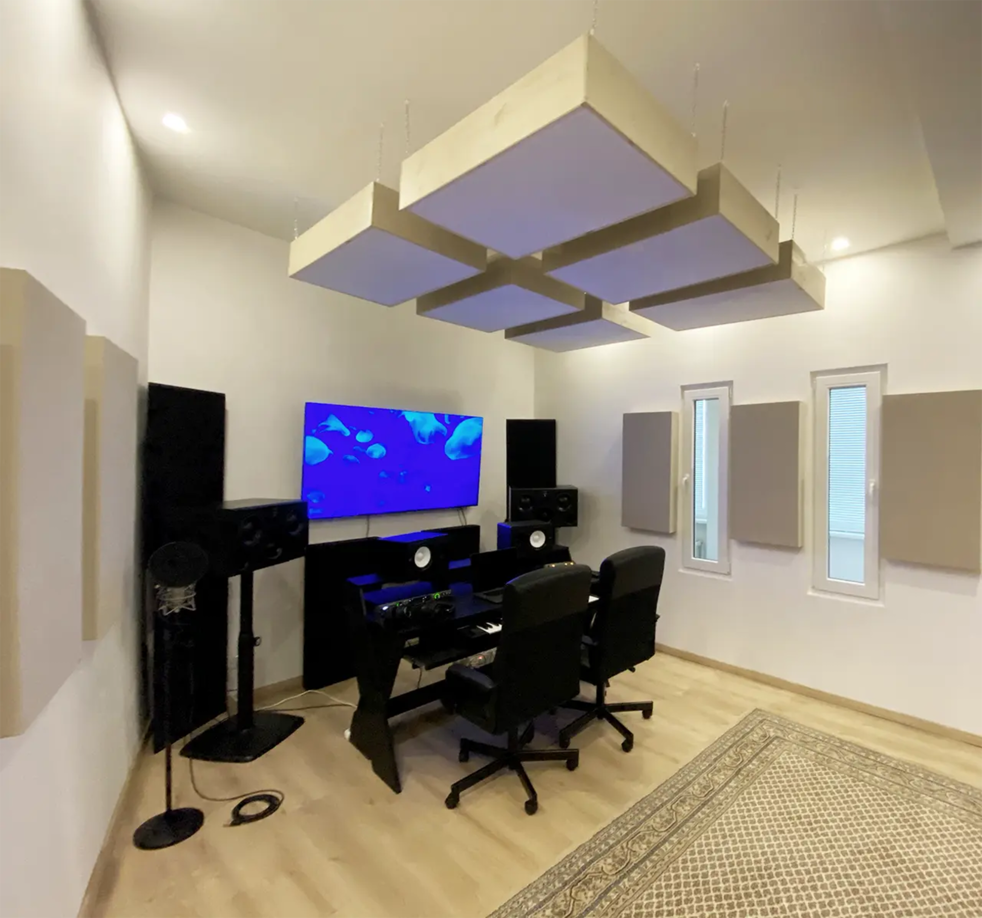 A modern recording studio with soundproofing panels, a mixing desk, two black chairs, and a large screen displaying a blue underwater scene.