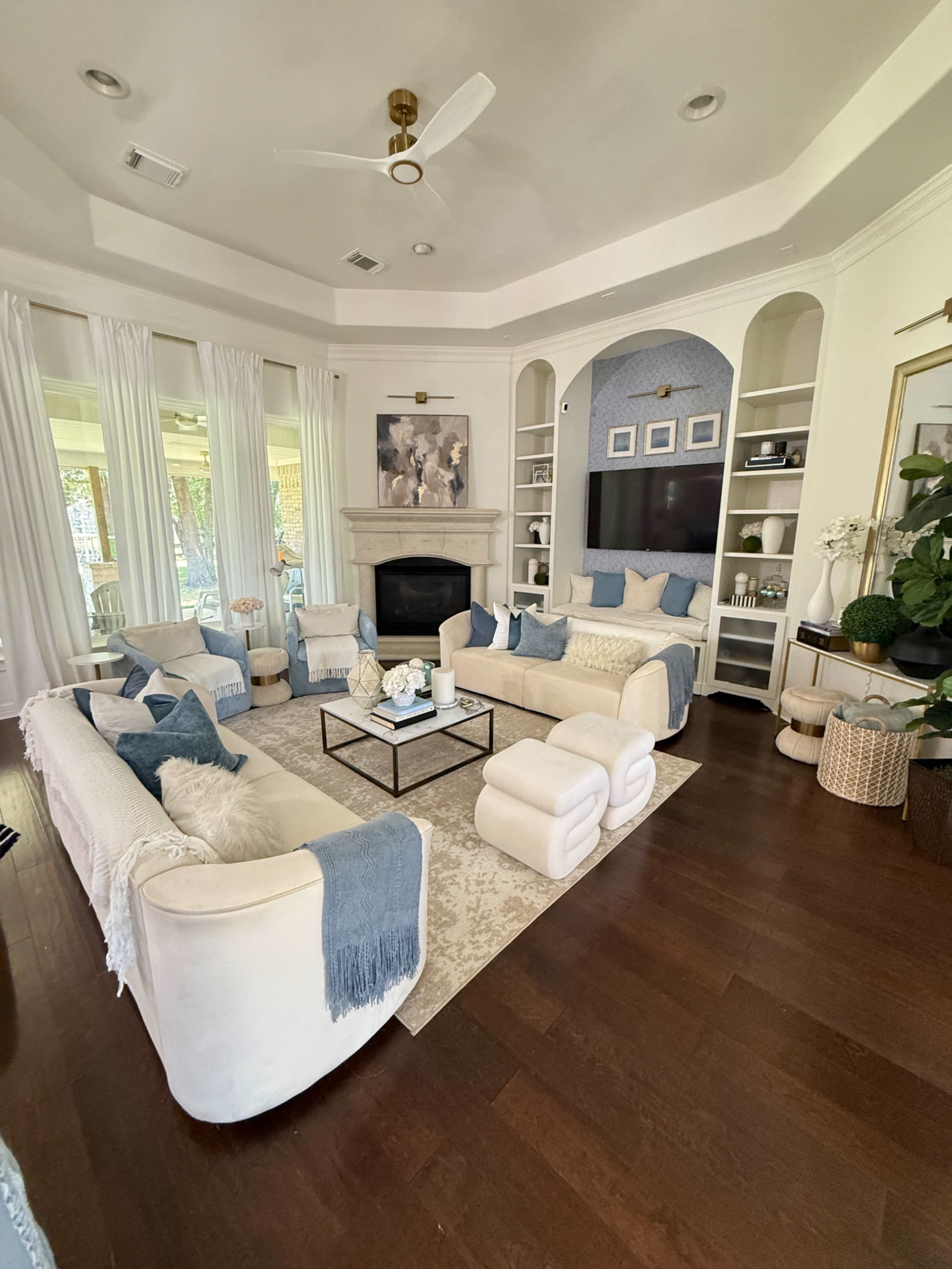 The image shows a neatly arranged living room with light-colored furniture, a central coffee table, and a stone fireplace surrounded by built-in shelves.