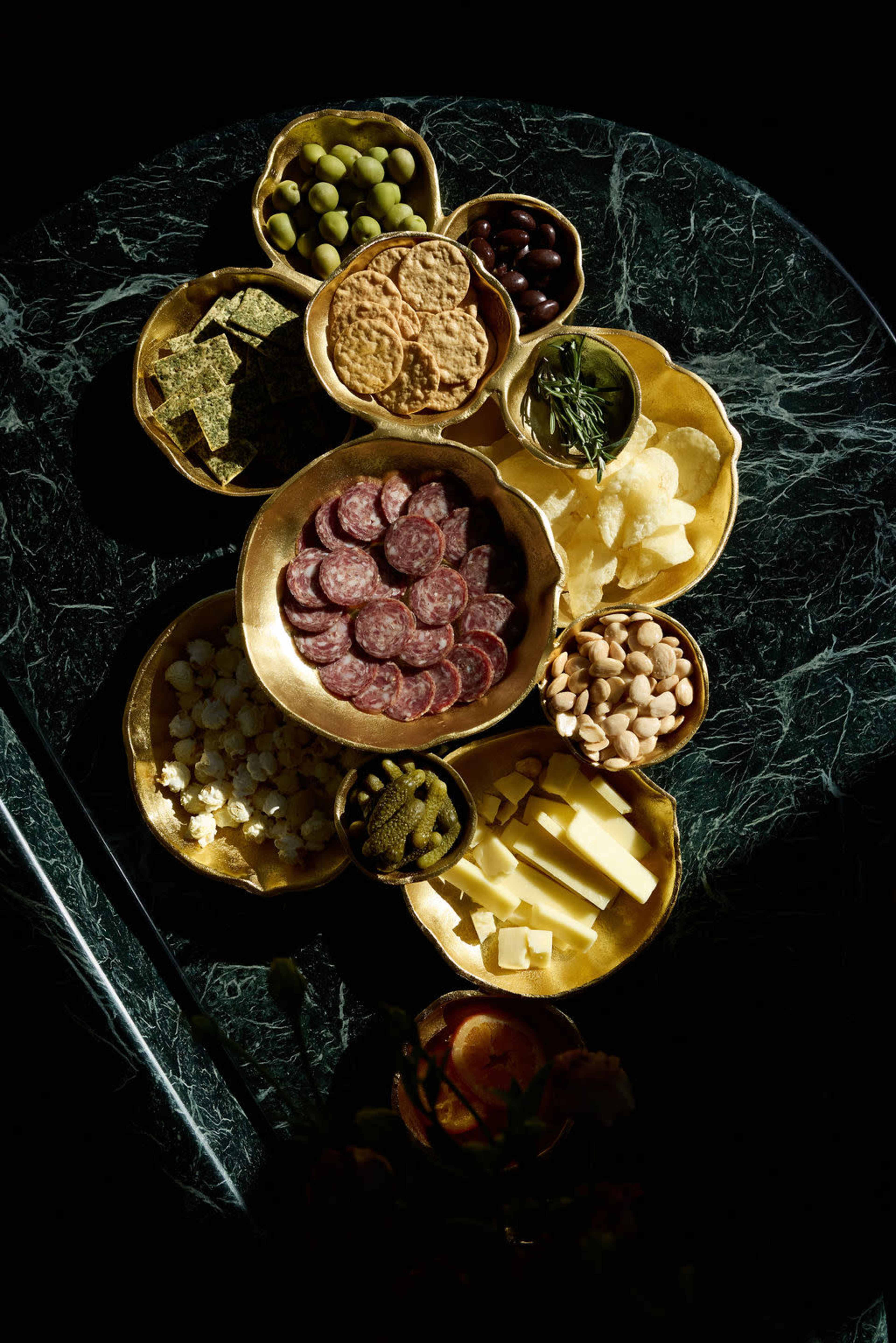 A variety of snacks, including meats, cheeses, olives, and crackers, are arranged in gold bowls on a dark marble surface.