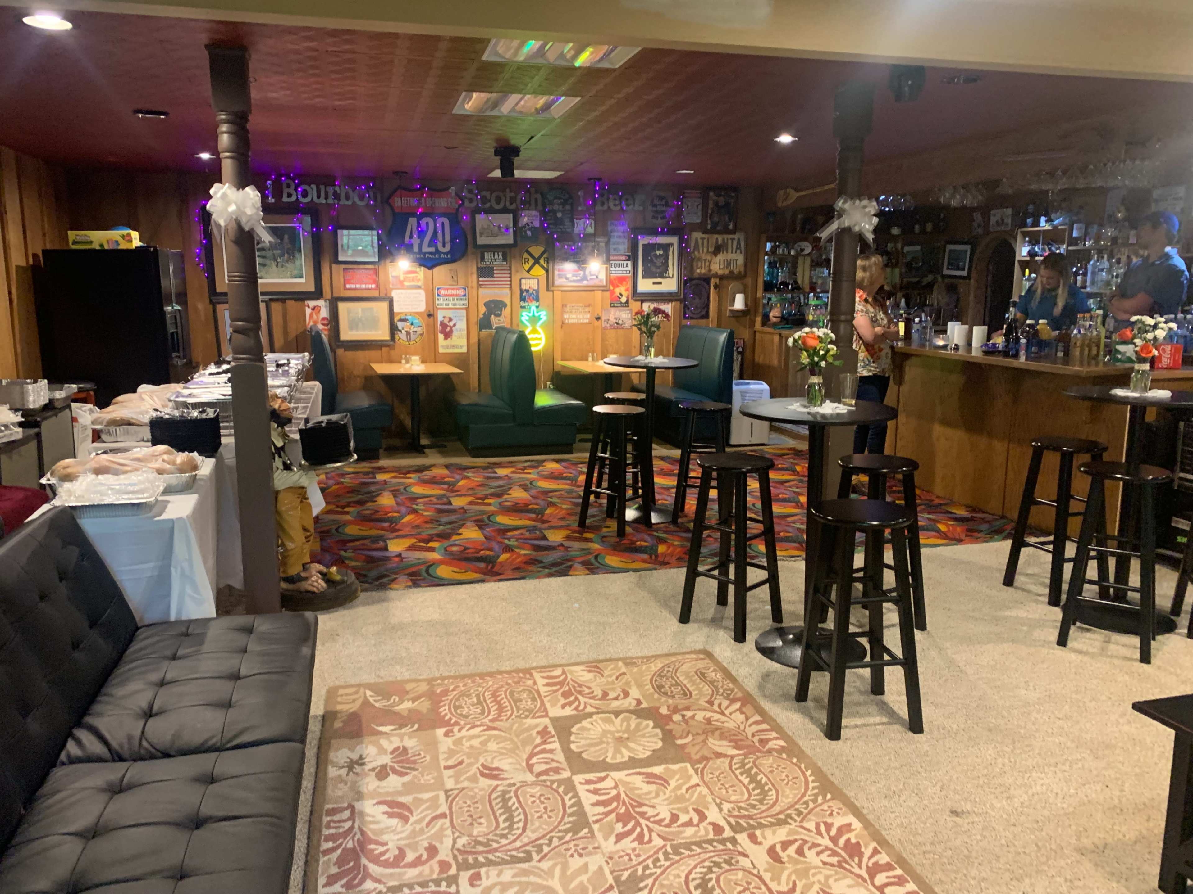 The image shows a lively bar area with a mix of seating, a decorated counter, and a buffet table set up against colorful carpeting.