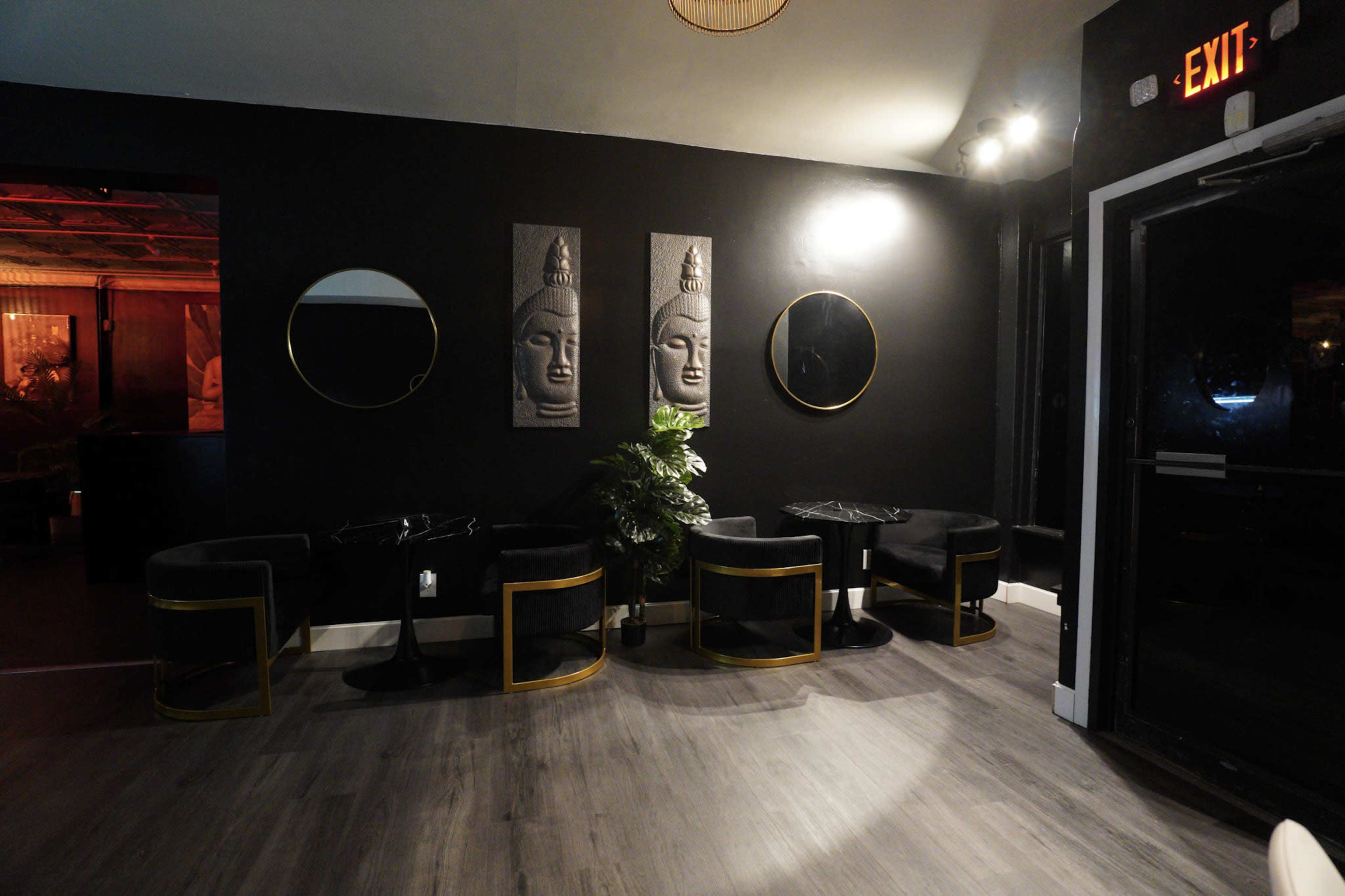 The interior of a dimly lit lounge features black walls, two round mirrors, and decorative Buddha wall art flanked by gold-accented seating and a small plant.