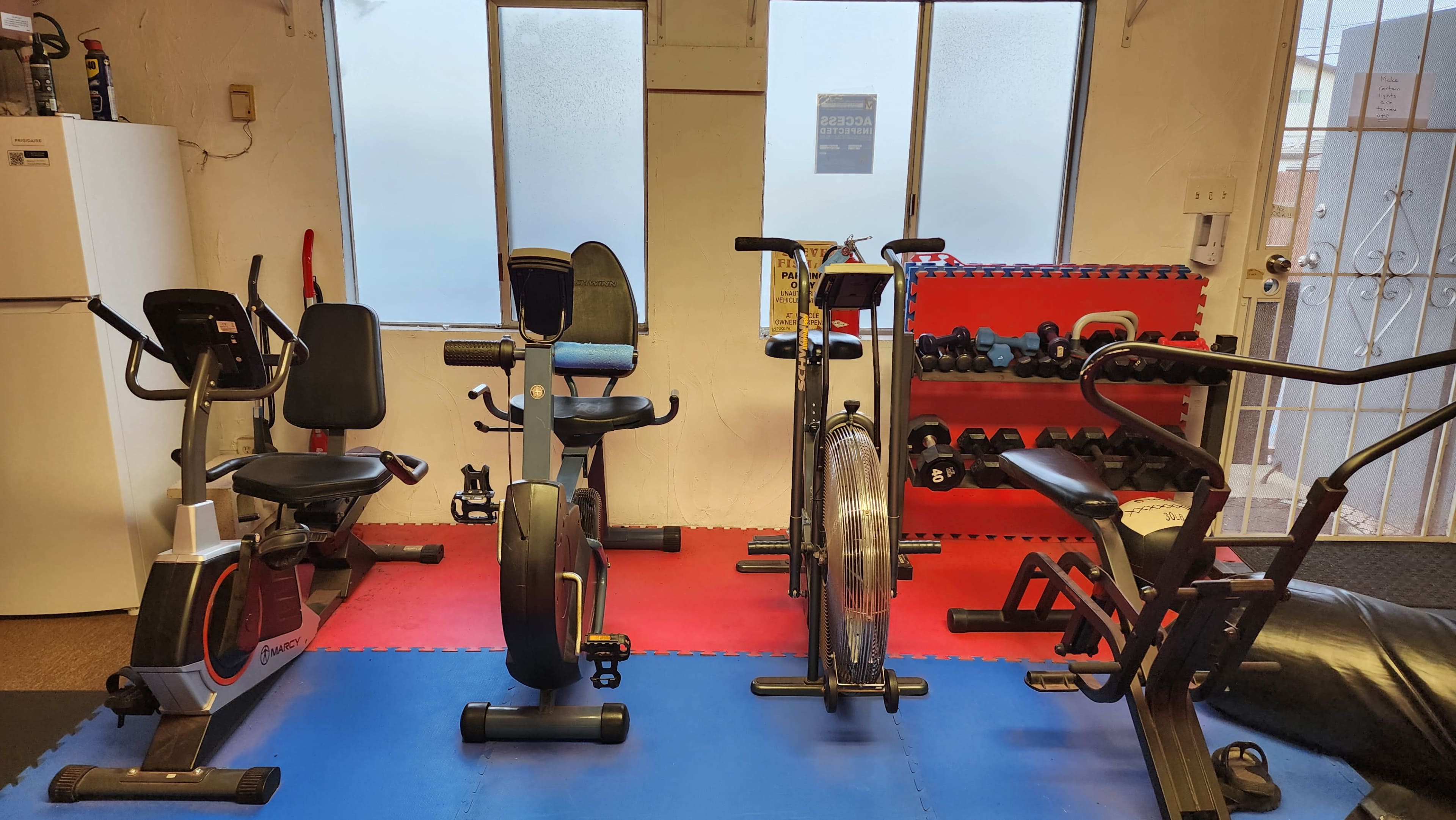 The image shows a small home gym setup featuring various exercise equipment, including a stationary bike, an air bike, and a weight bench, with free weights on a rack against the wall.