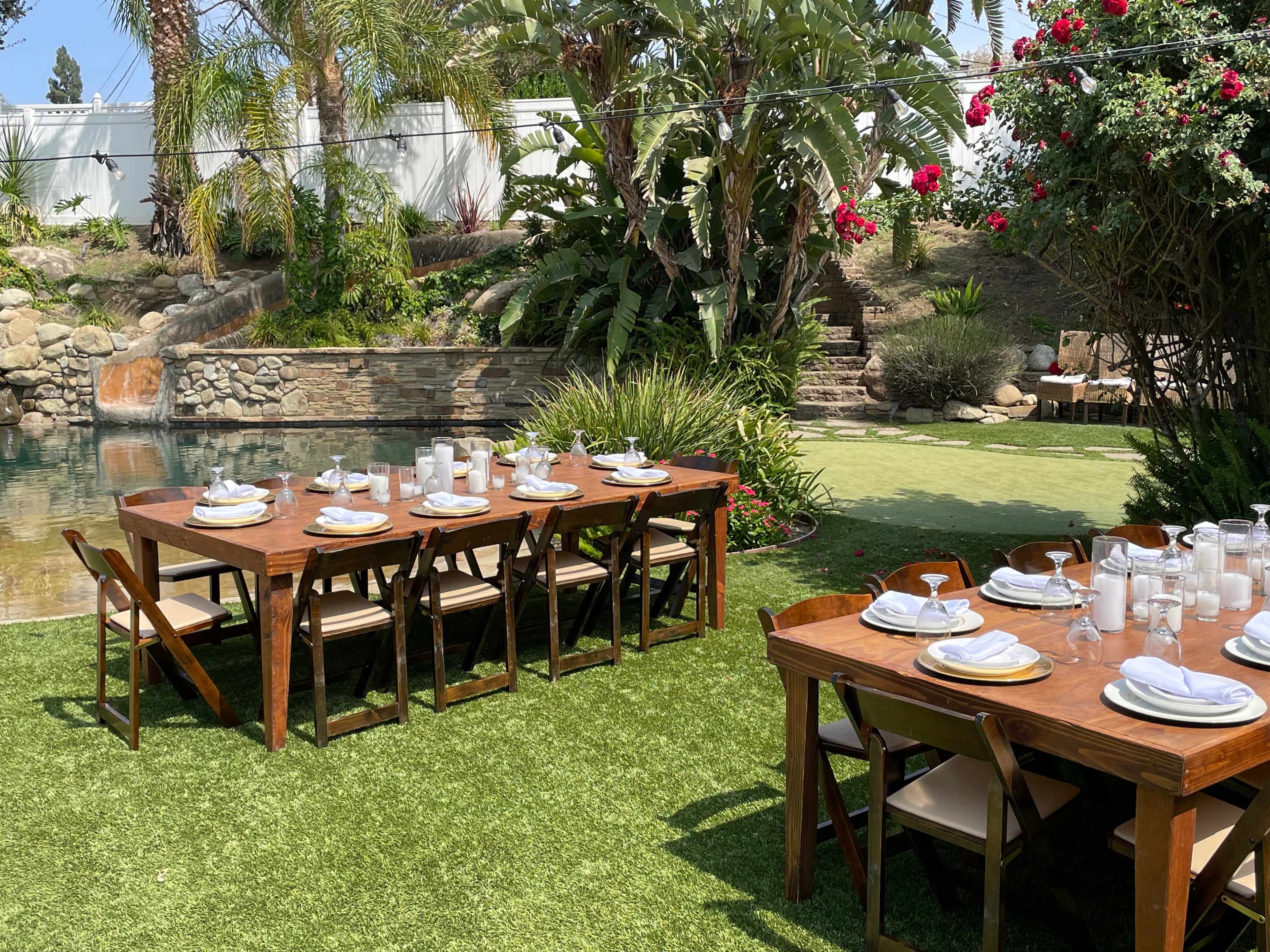 A landscaped backyard features two wooden dining tables set with tableware beside a swimming pool and surrounded by tropical plants.