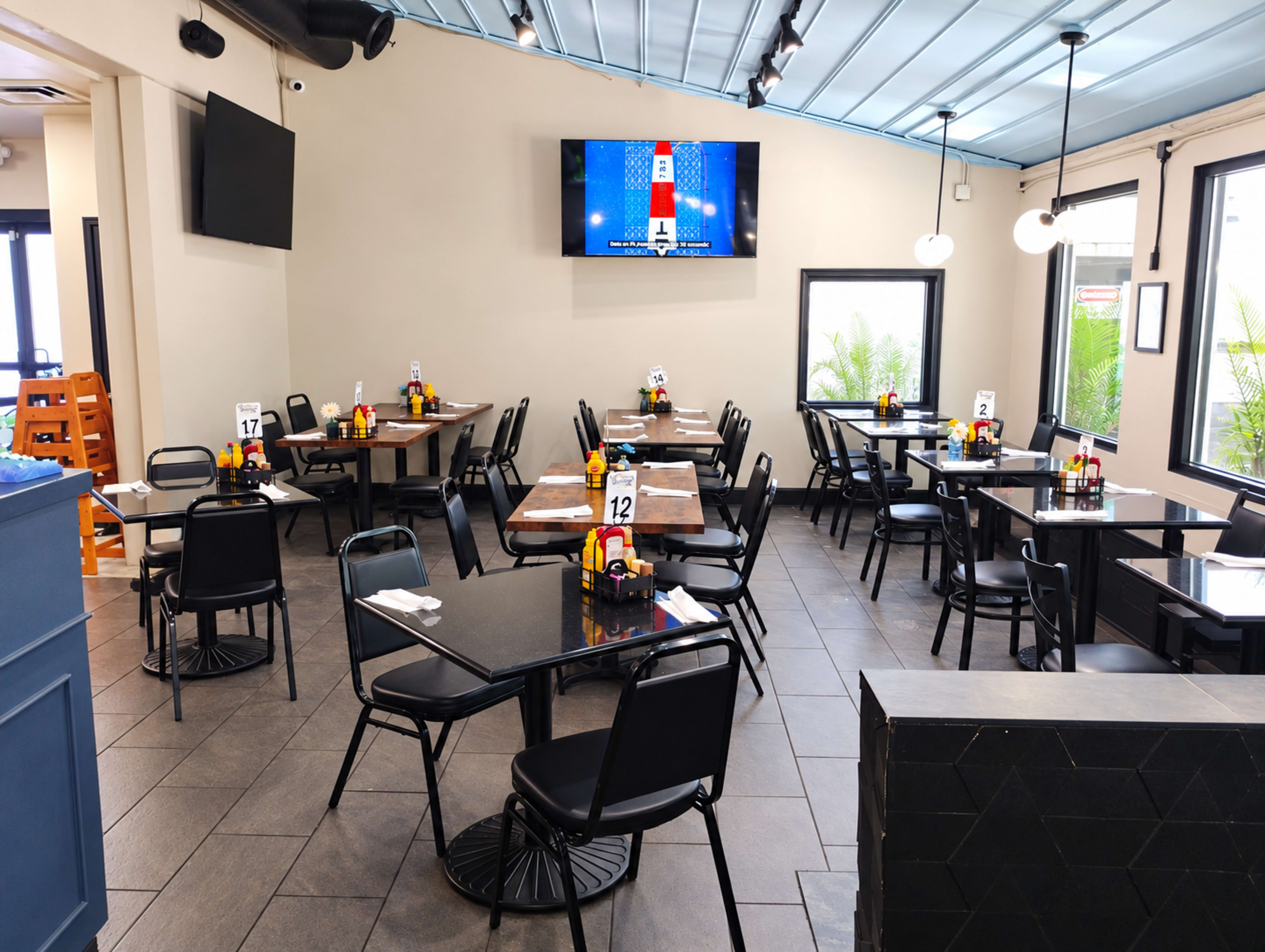 The image shows a modern restaurant interior with empty tables, black chairs, menu items on each table, and a large wall-mounted television displaying content.