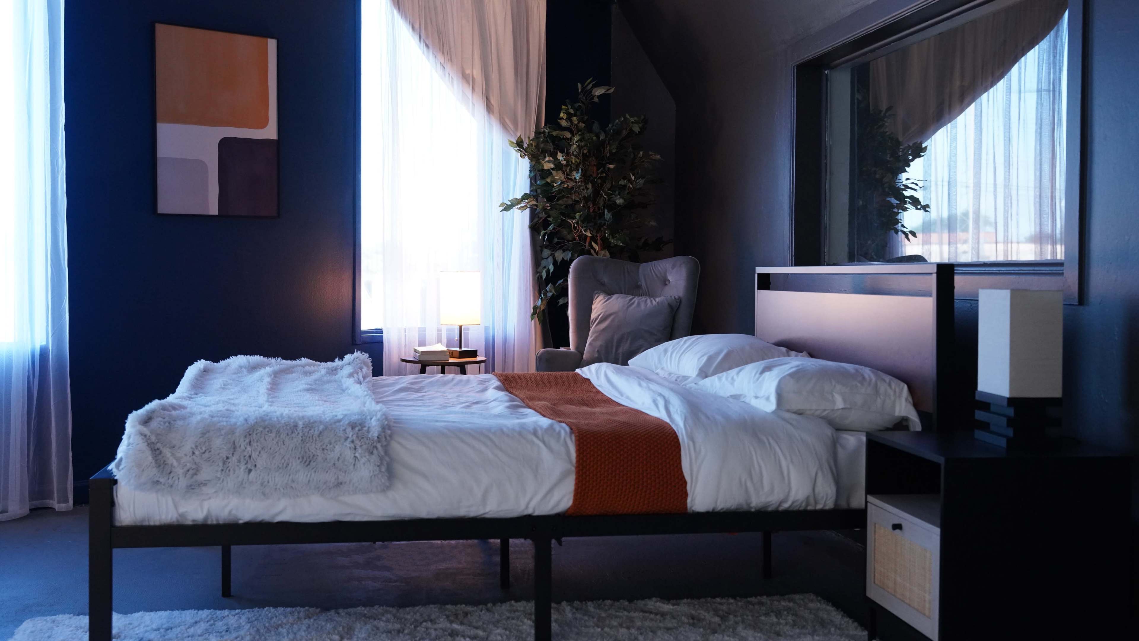 A neatly made bed in a modern bedroom featuring dark walls, large windows with sheer curtains, and a decorative plant near the corner.