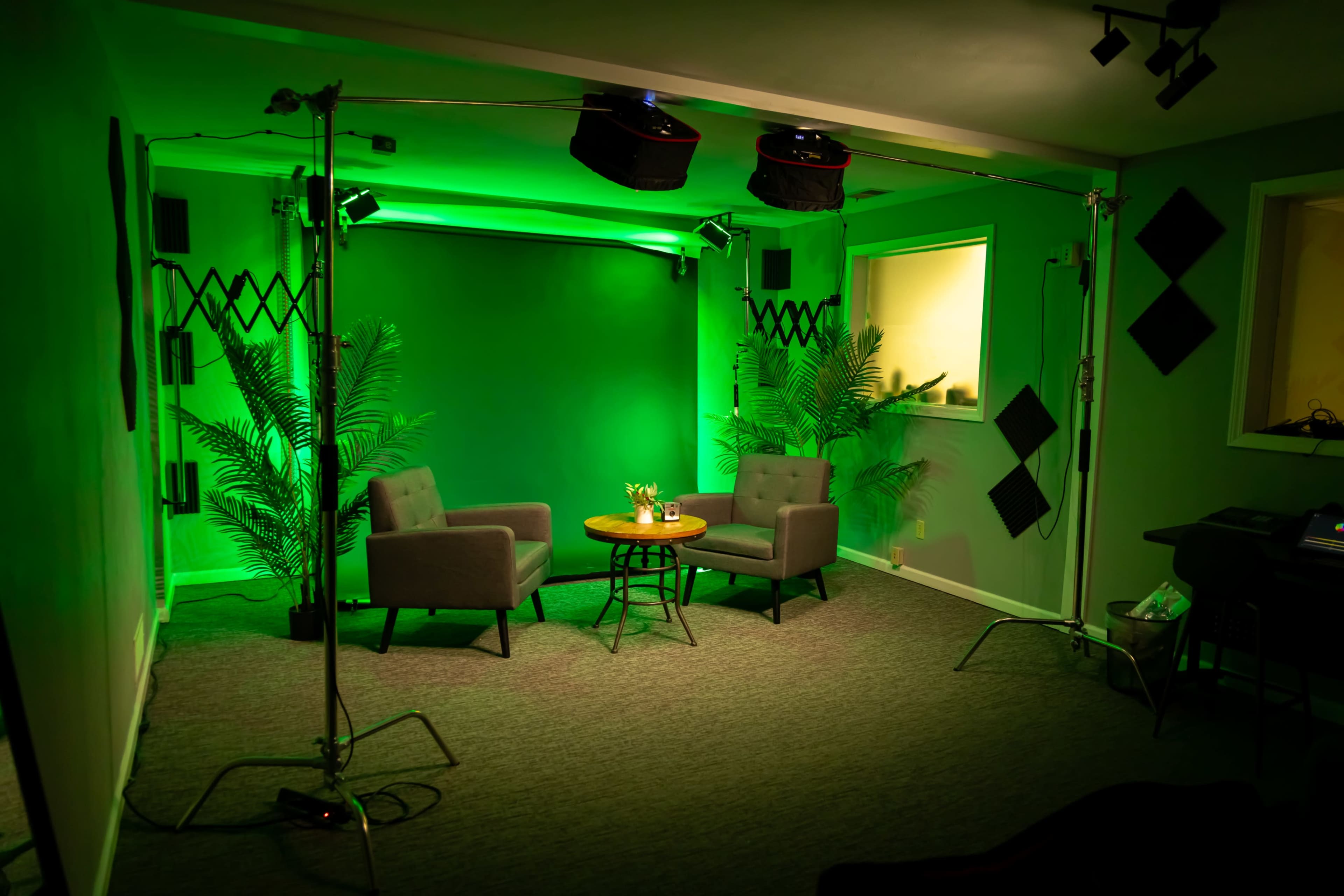 The image shows a small studio setup with two gray chairs, a wooden table, and green lighting, surrounded by potted plants and microphone equipment.