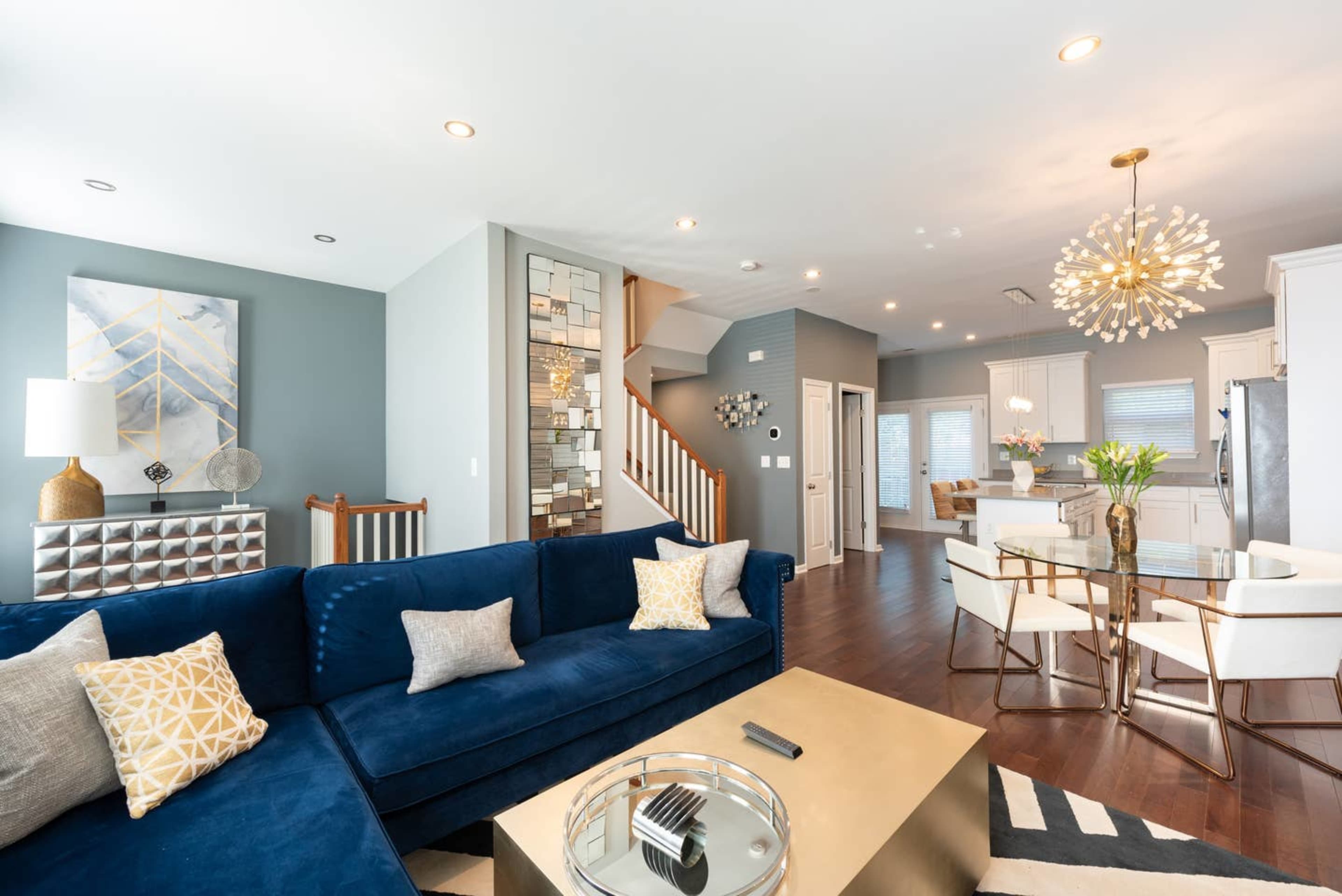A modern living room featuring a blue velvet sofa, a gold coffee table, and a dining area with white chairs, illuminated by a stylish light fixture.