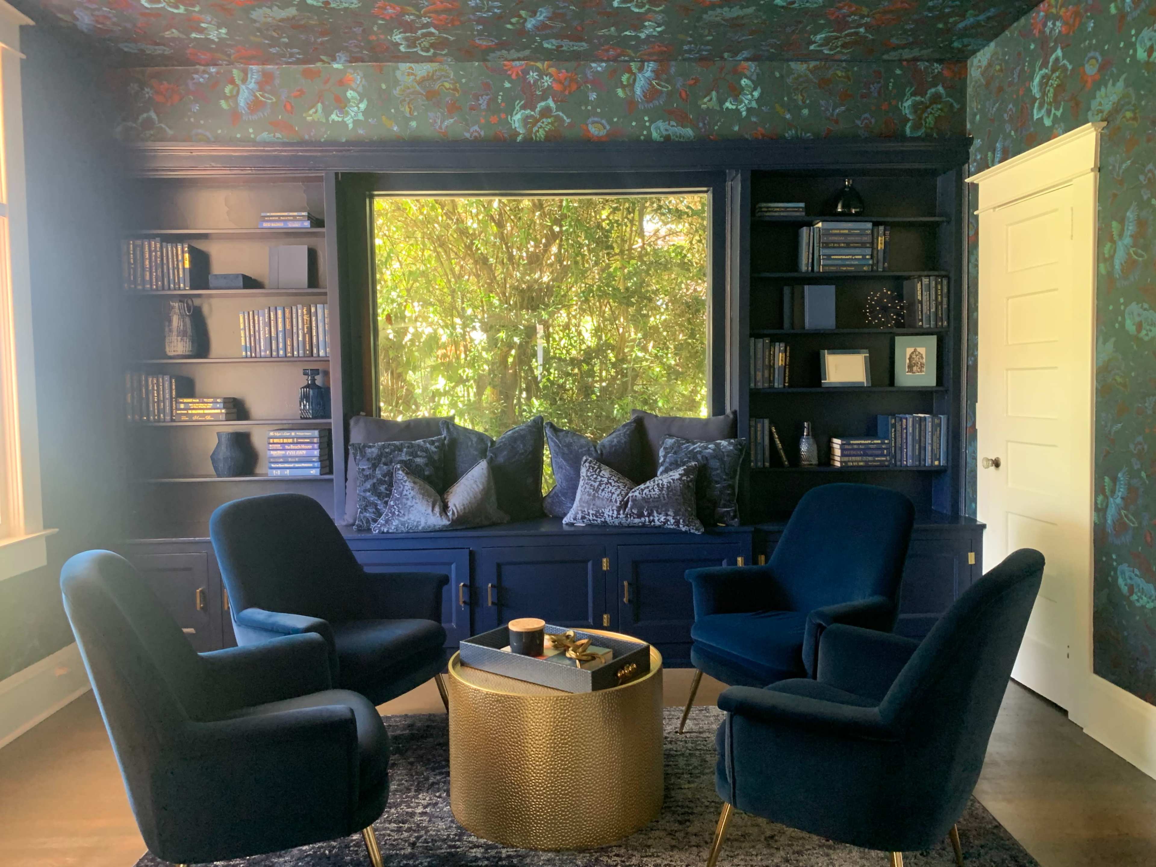 A cozy seating area features four blue chairs arranged around a metallic gold coffee table, with bookshelves and a large window adorned with leafy views in the background.
