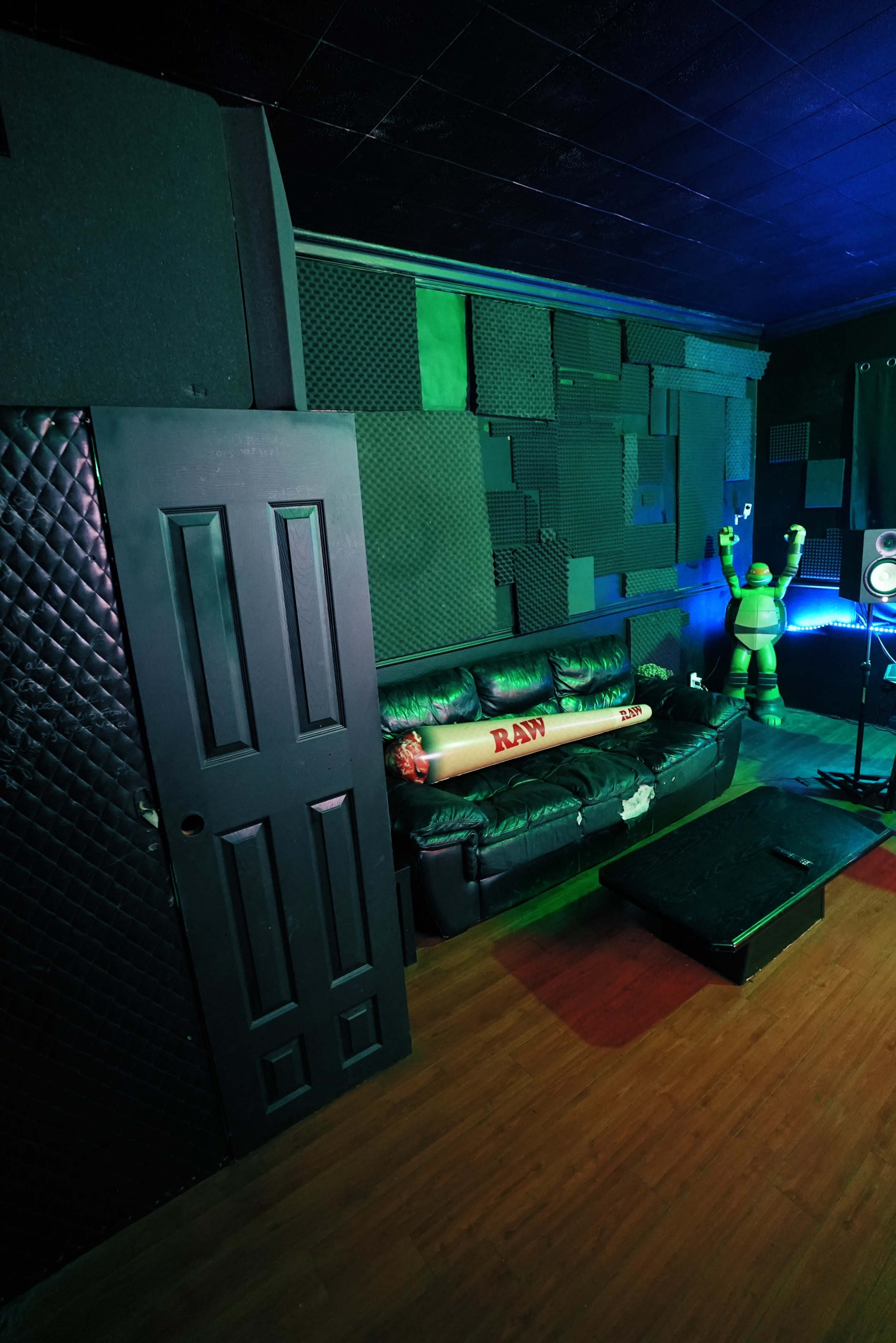 The image shows a sound studio with a black door, a green-lit corner featuring a couch, and soundproofing panels on the walls.