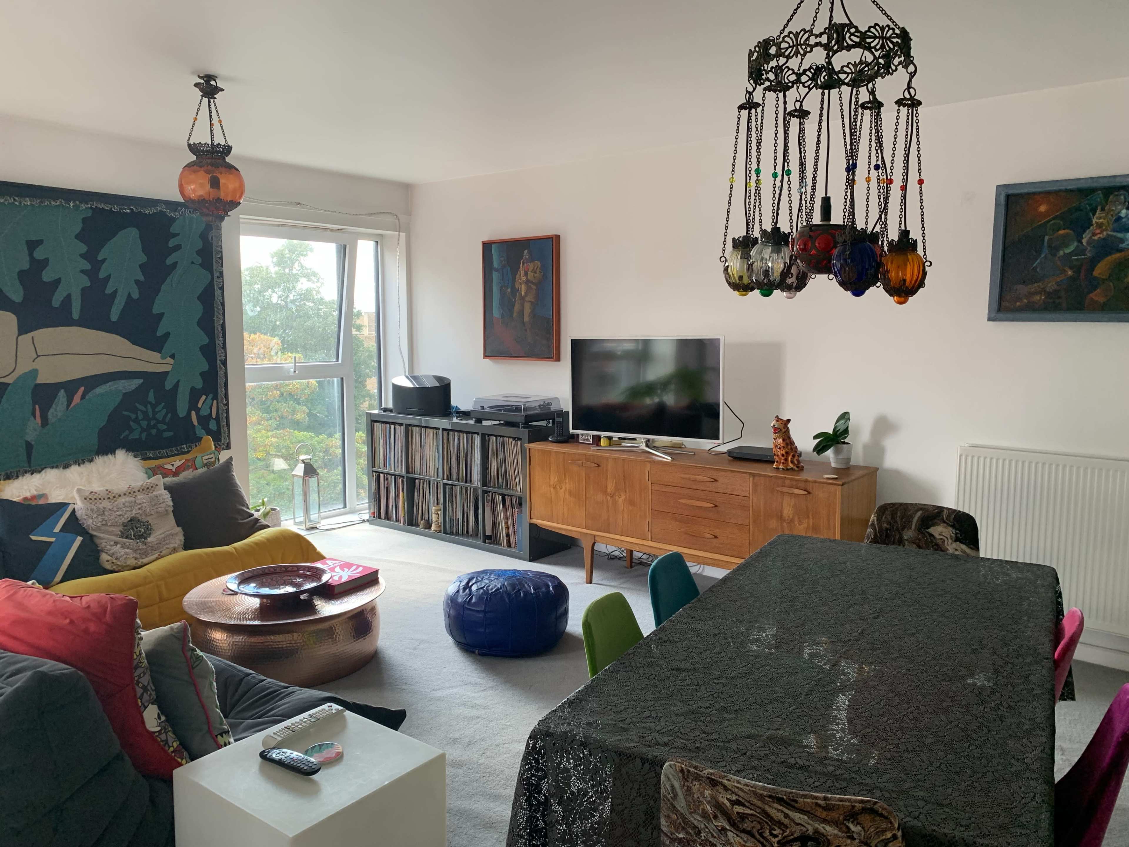 A living room features a large dining table, colorful chairs, a television on a wooden cabinet, and decorative lighting hanging from the ceiling.