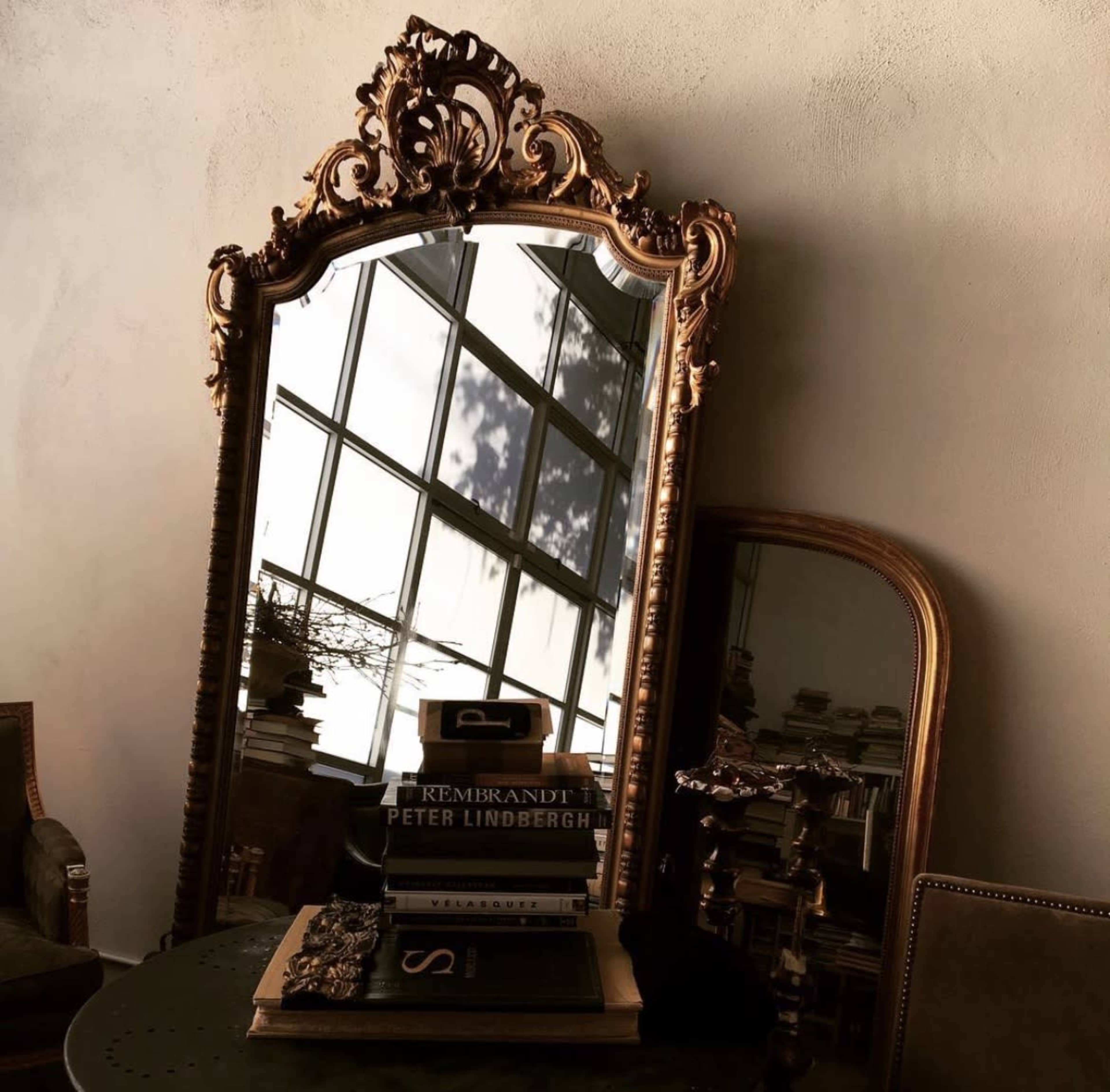 An ornate, vintage mirror with a decorative frame placed against a wall, reflecting a window and a stack of books on a round table.