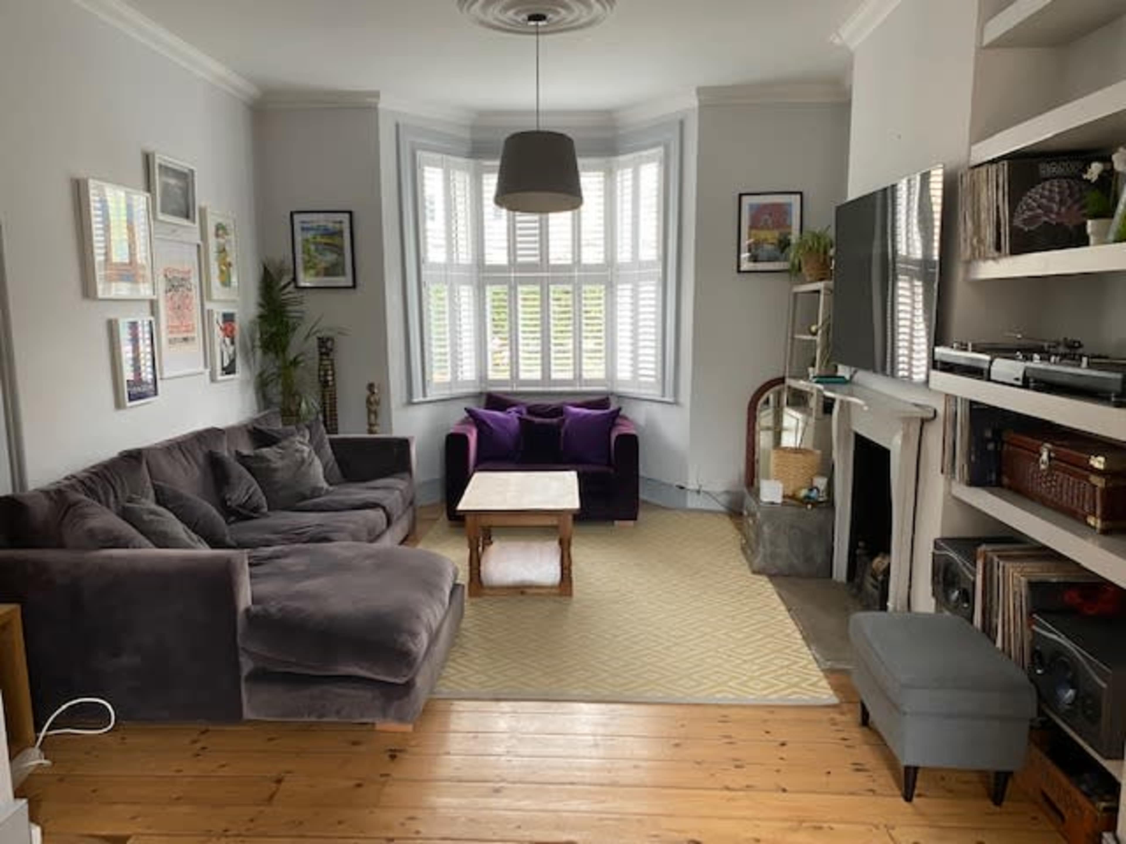 The living room features a gray sectional sofa, two purple armchairs, a hardwood floor, and a large bay window with white shutters.