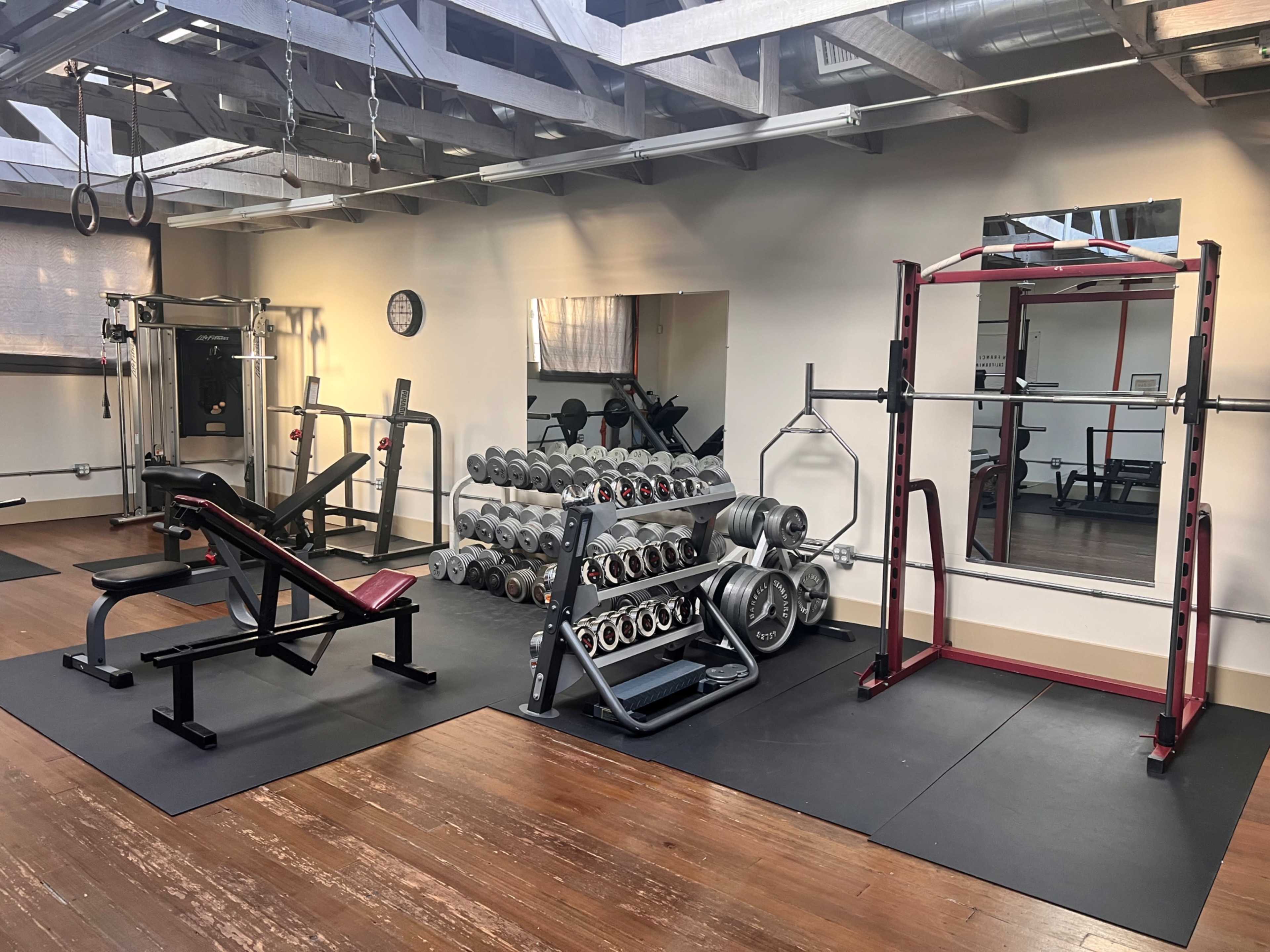 A gym area features a weight bench, a squat rack, a dumbbell rack, and various exercise equipment arranged on a wooden floor.