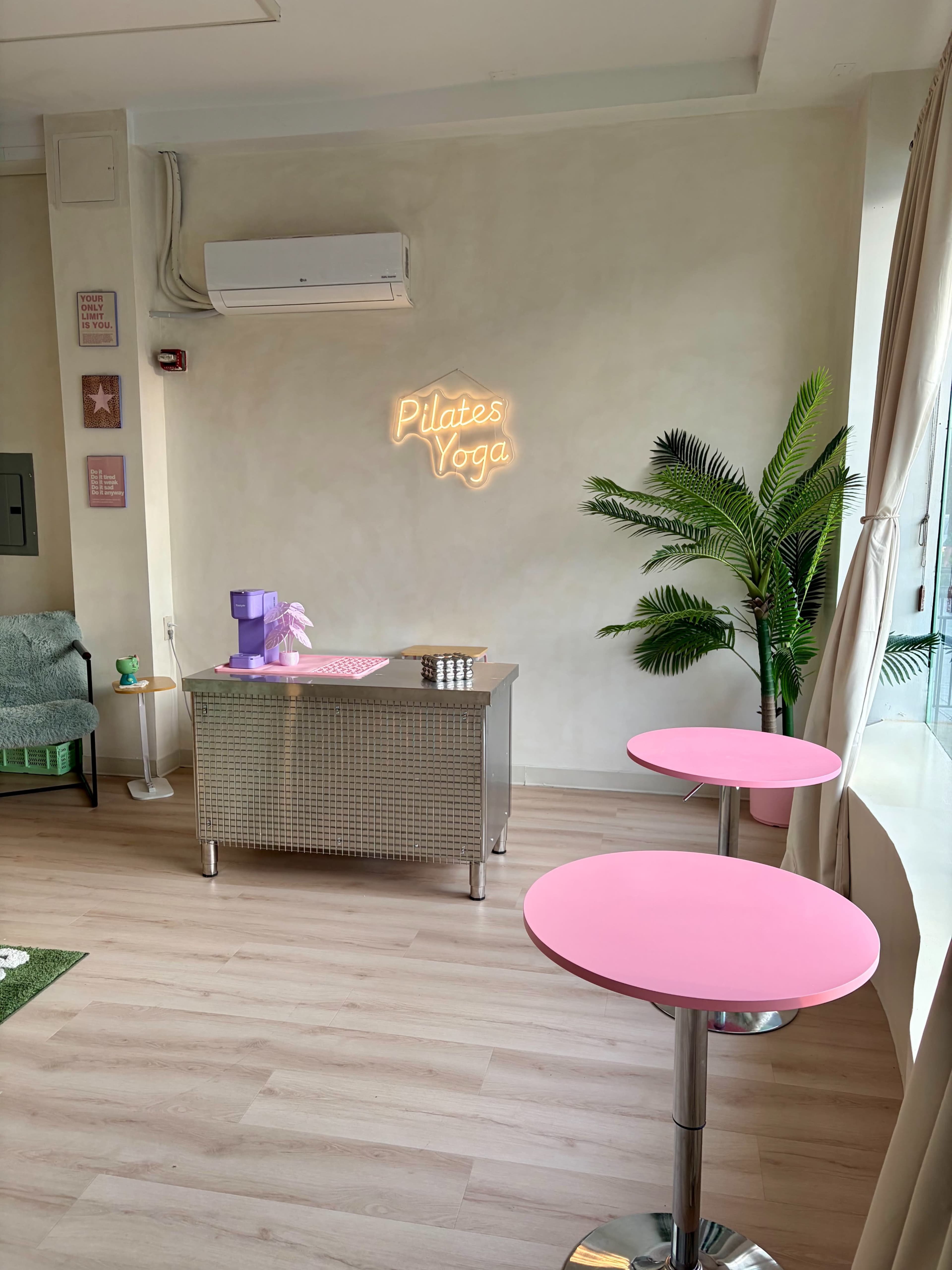 The image shows a bright, minimalistic interior with pink tables, a metallic desk, and a neon sign that reads "Pilates Yoga," accompanied by a potted plant.