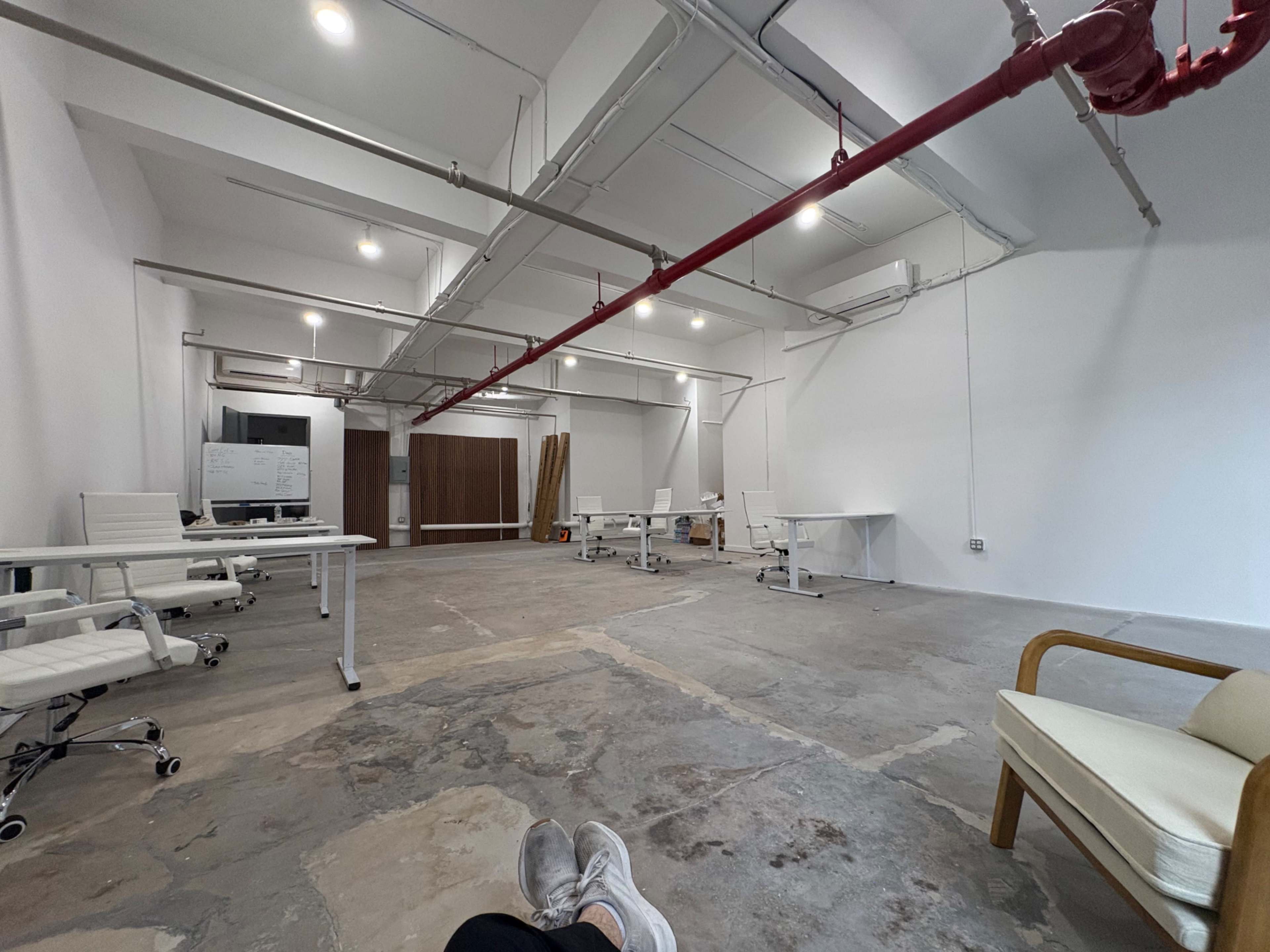The image shows a spacious, partially furnished office with white walls, exposed piping, and several empty desks and chairs.