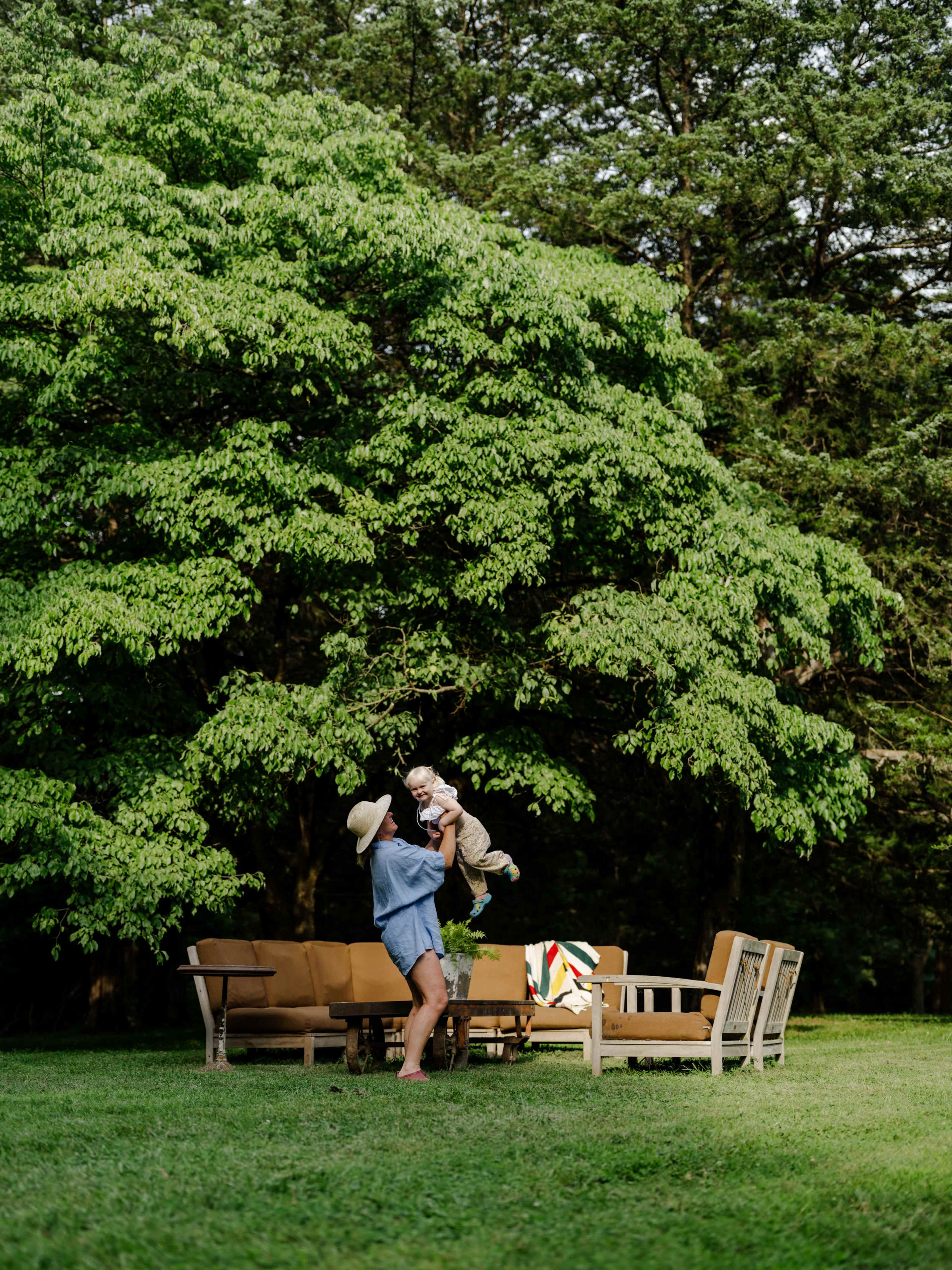 A person lifts a child in the air beside a seating area under a large tree in a grassy outdoor space.