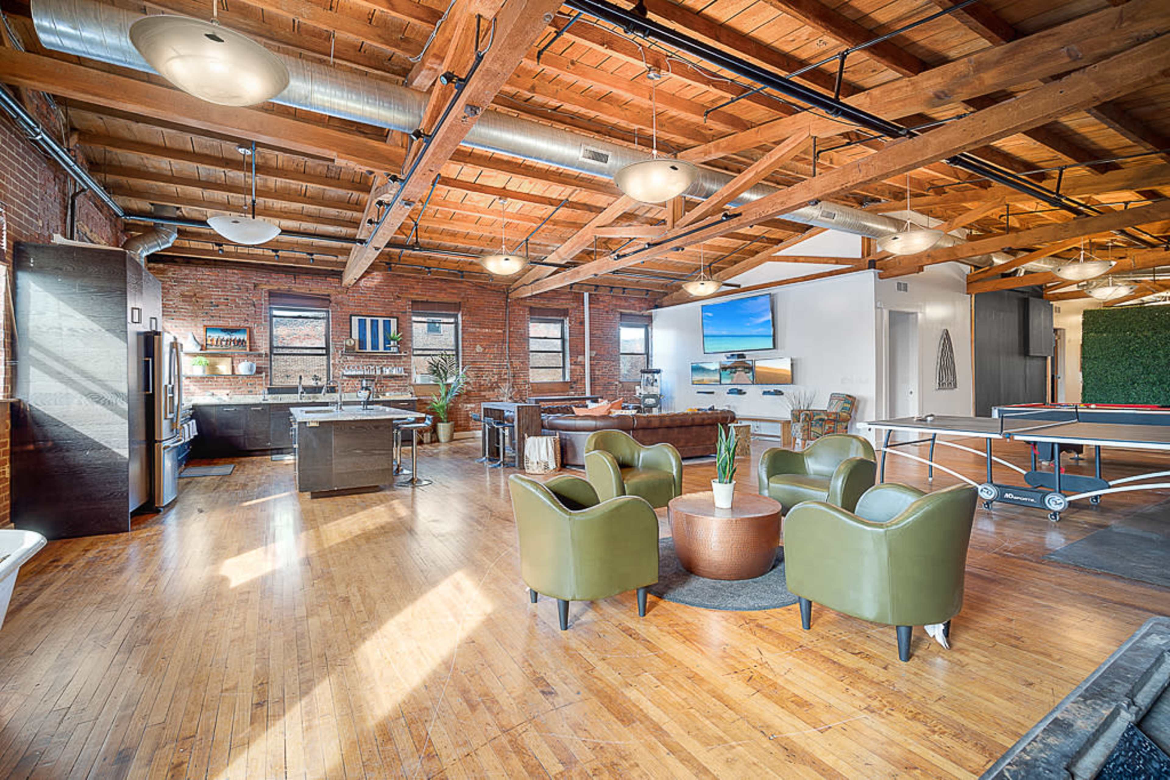 A spacious loft with wooden beams and floors, featuring a seating area with green chairs, a kitchen, and a games table.