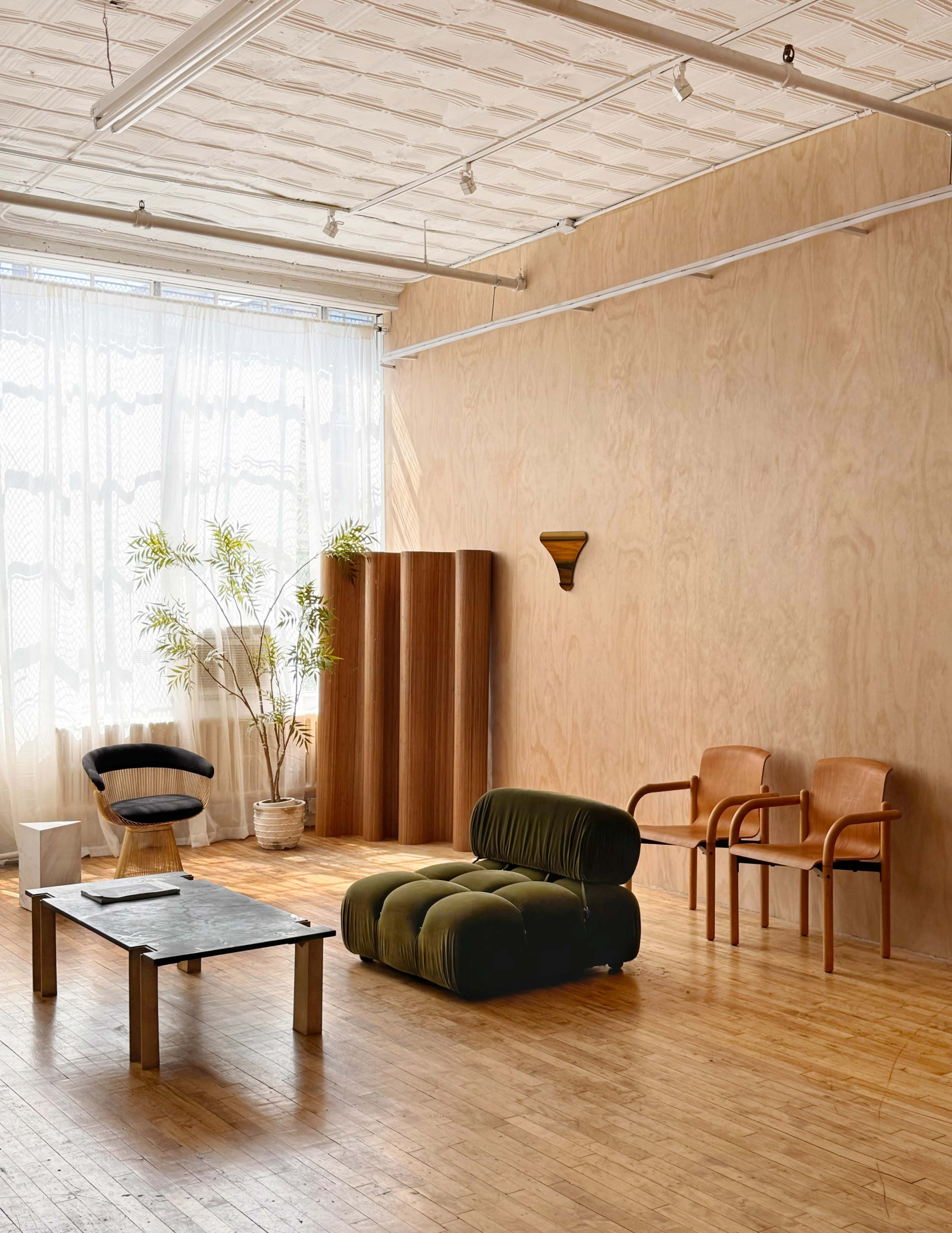 The image shows a minimalist room with a wooden floor, a large window with sheer curtains, a green upholstered chair, a black coffee table, two leather chairs, and a potted plant.