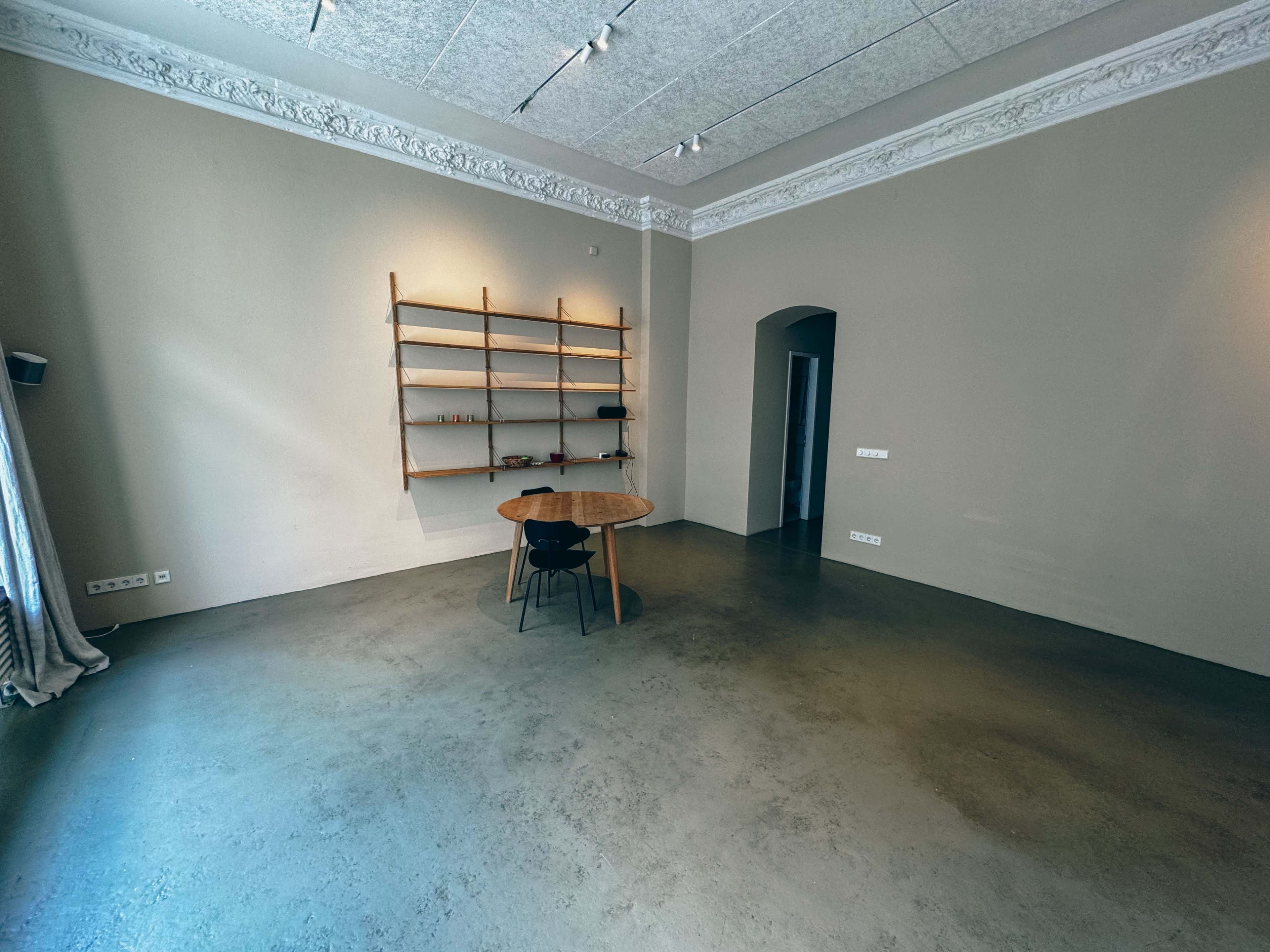 The image shows a spacious room featuring a round wooden table, a minimalist wall shelf with items, and a doorway leading to another room.