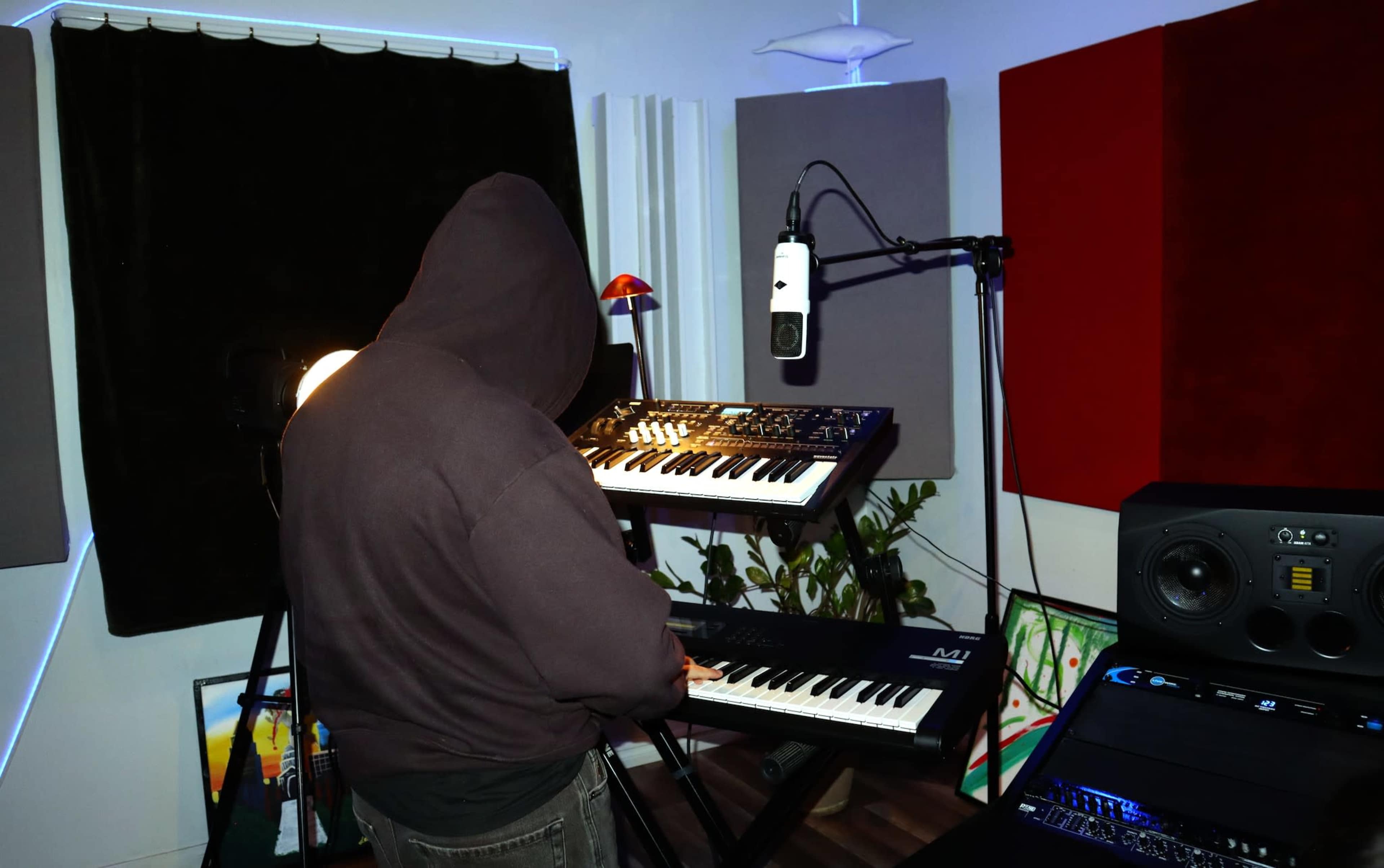 A person wearing a hooded sweatshirt stands in a music studio, surrounded by synthesizers, a microphone, and decorative artwork.