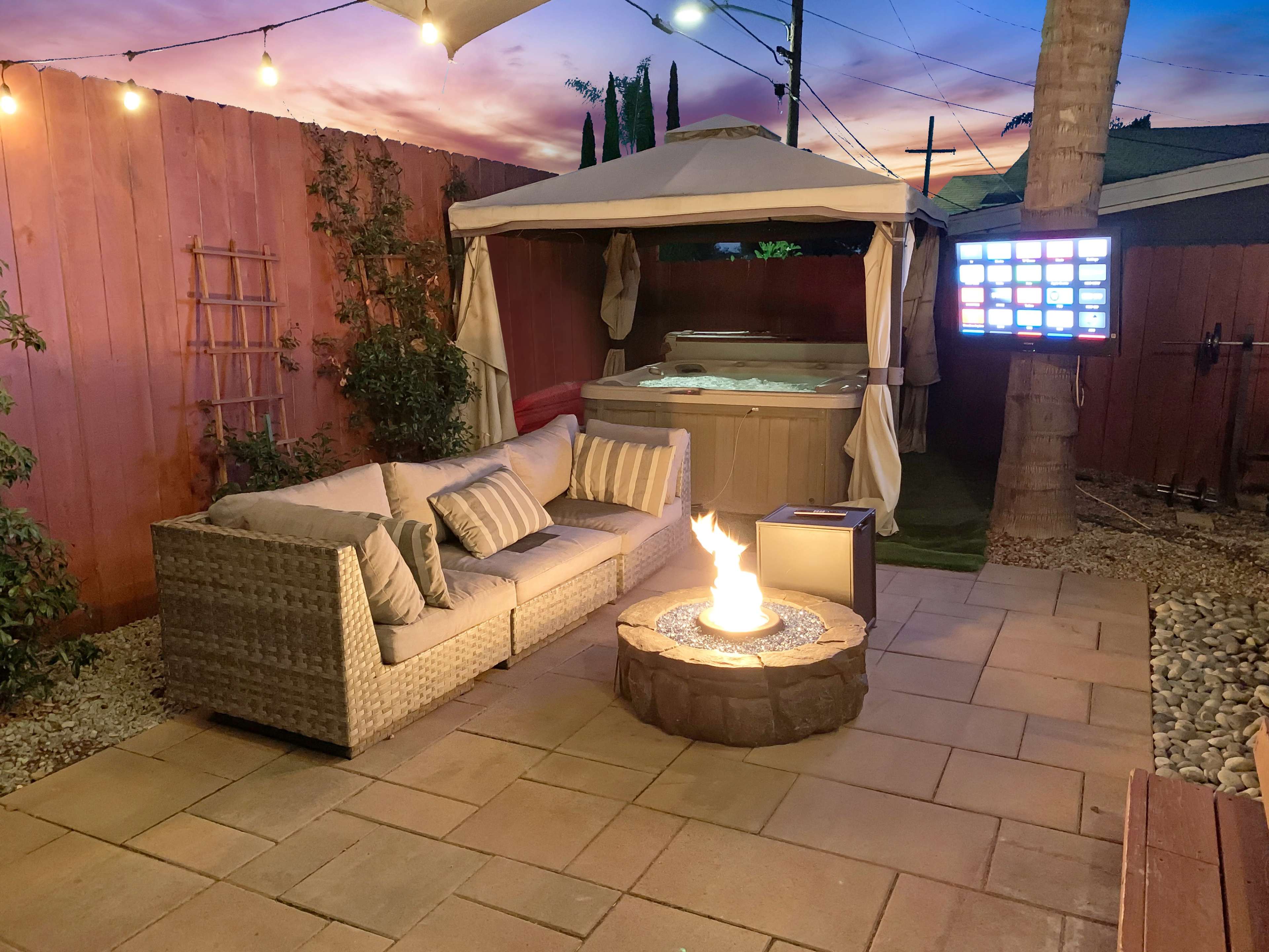 The scene features a cozy outdoor patio with a sectional sofa, a fire pit, a hot tub, and a television under a canopy, framed by wooden fences and string lights.