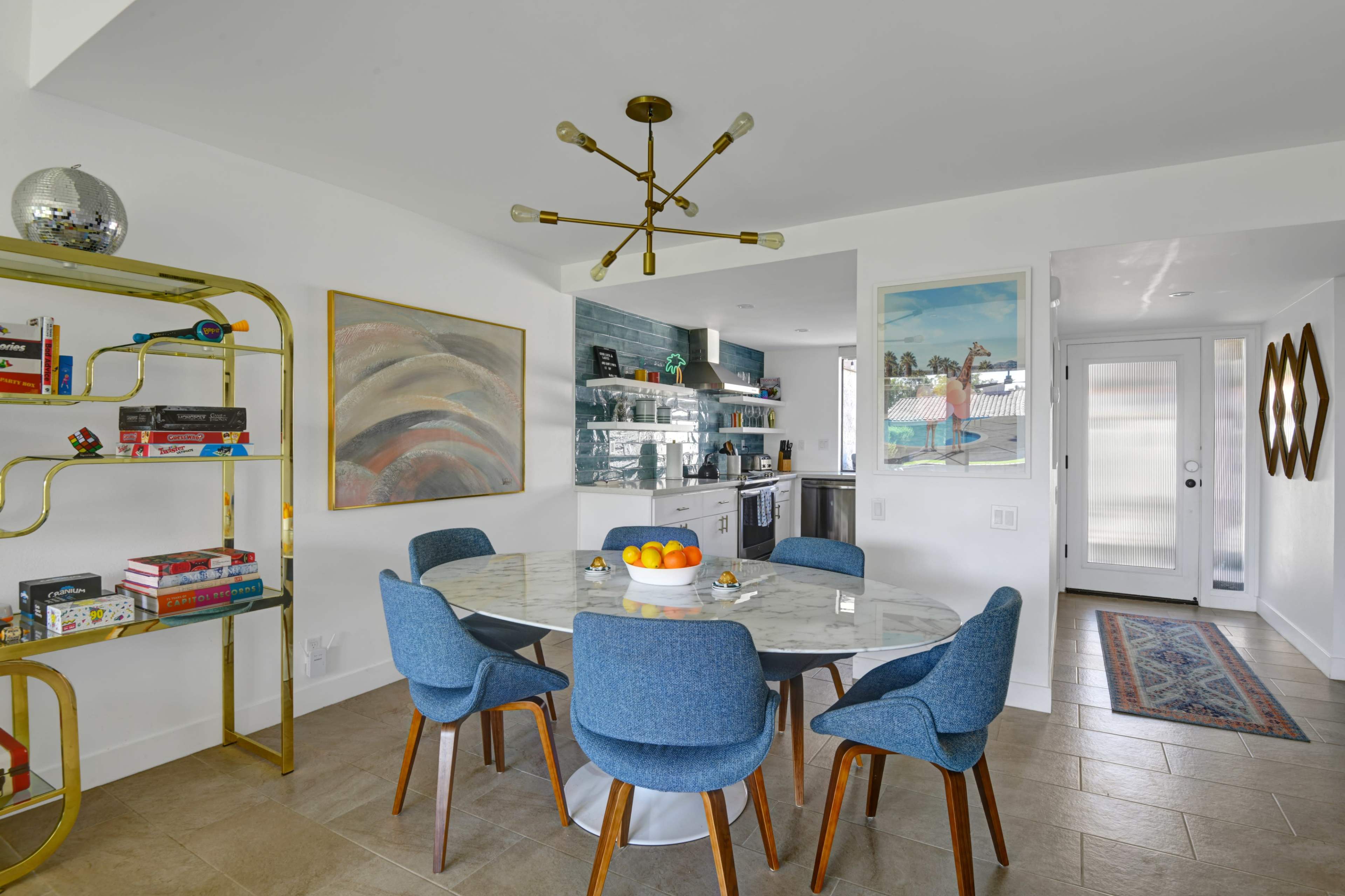 A modern dining area features a round table with blue upholstered chairs, a gold bookshelf filled with games and books, and a well-lit kitchen in the background.
