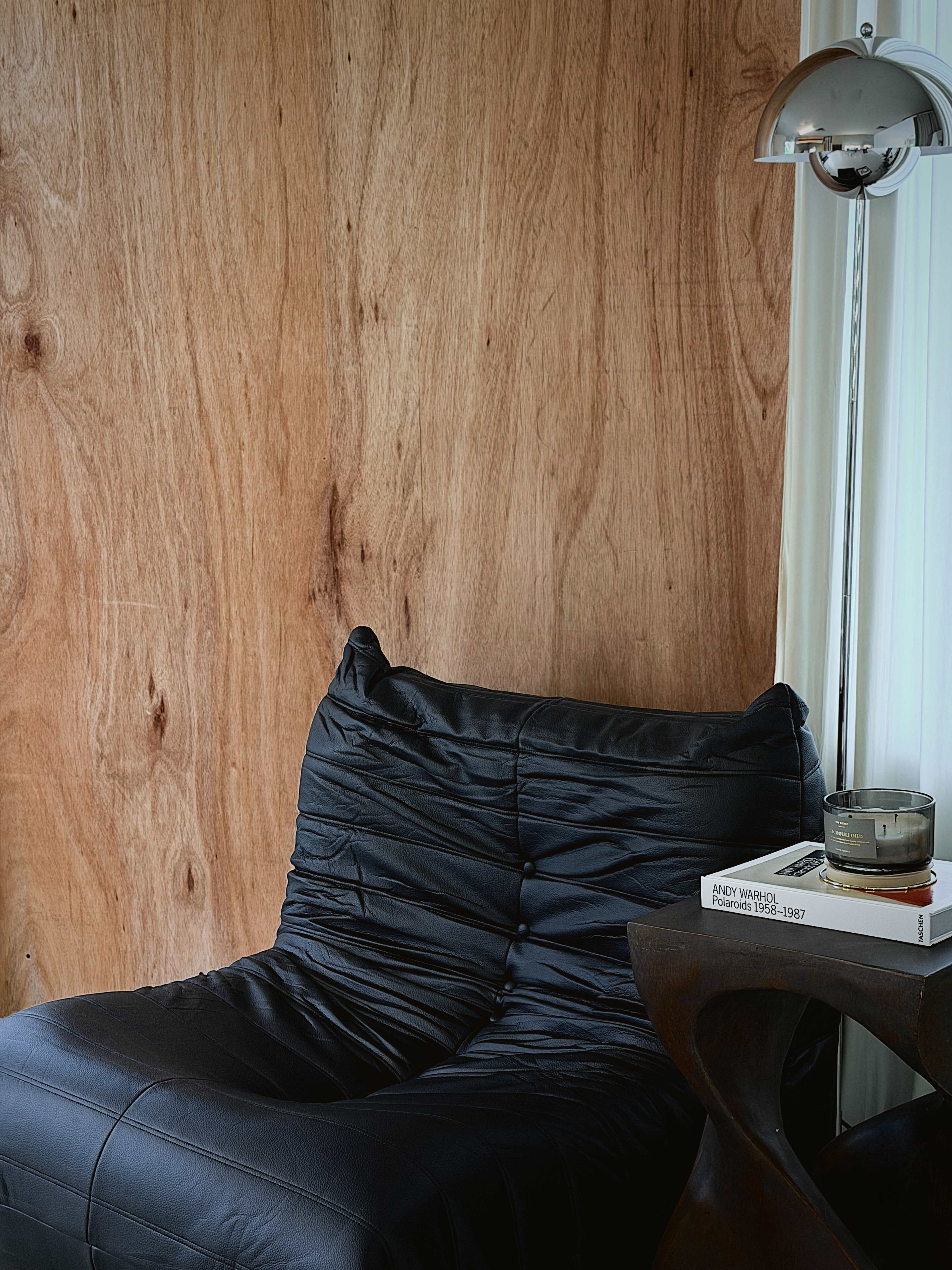 The image features a black leather chair positioned against a wooden wall, accompanied by a modern lamp and a stack of books on a side table.