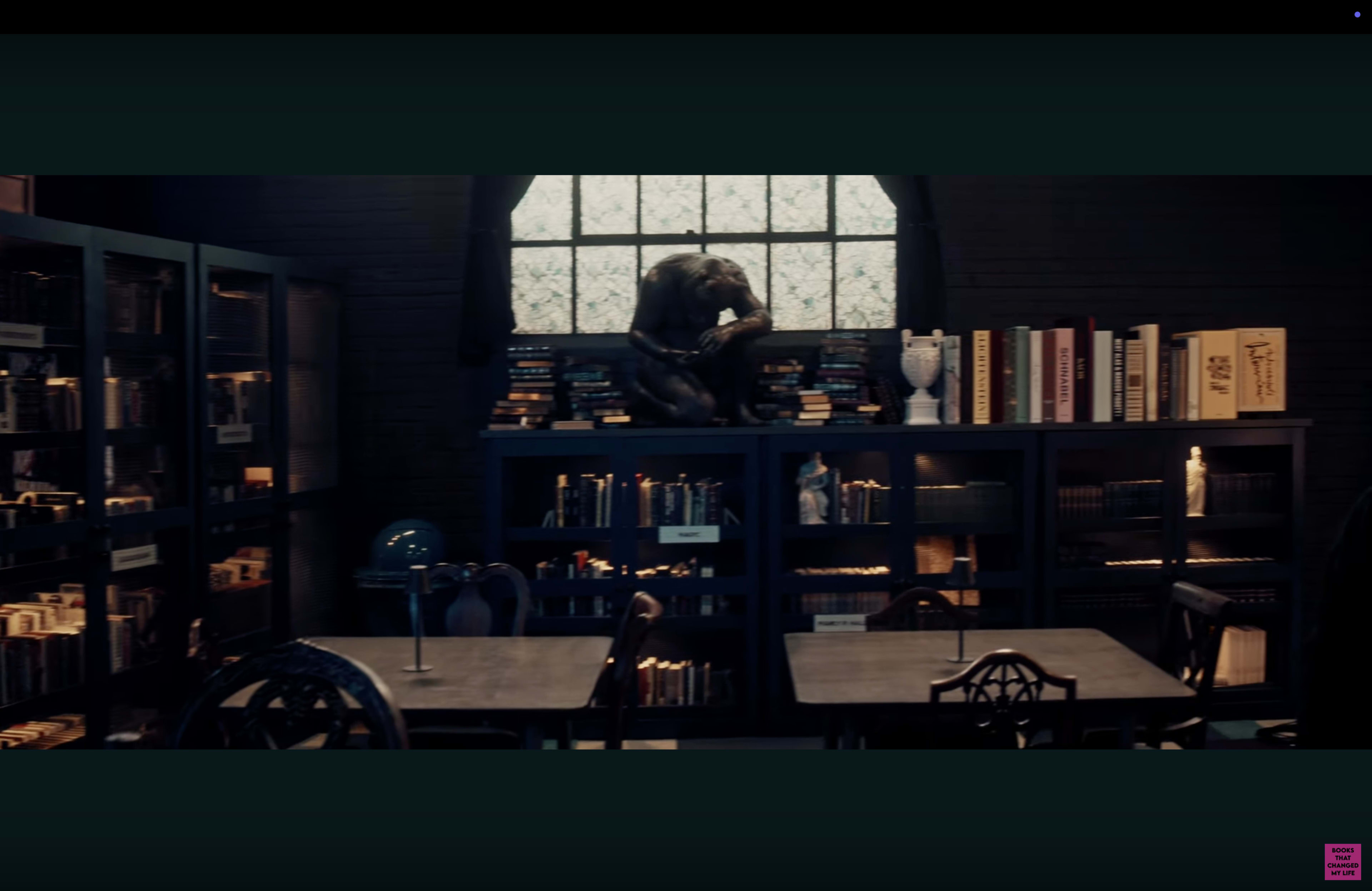 The image depicts a dimly lit library with dark wooden shelves filled with books, a sculpture on a shelf, and tables with chairs arranged in the foreground.
