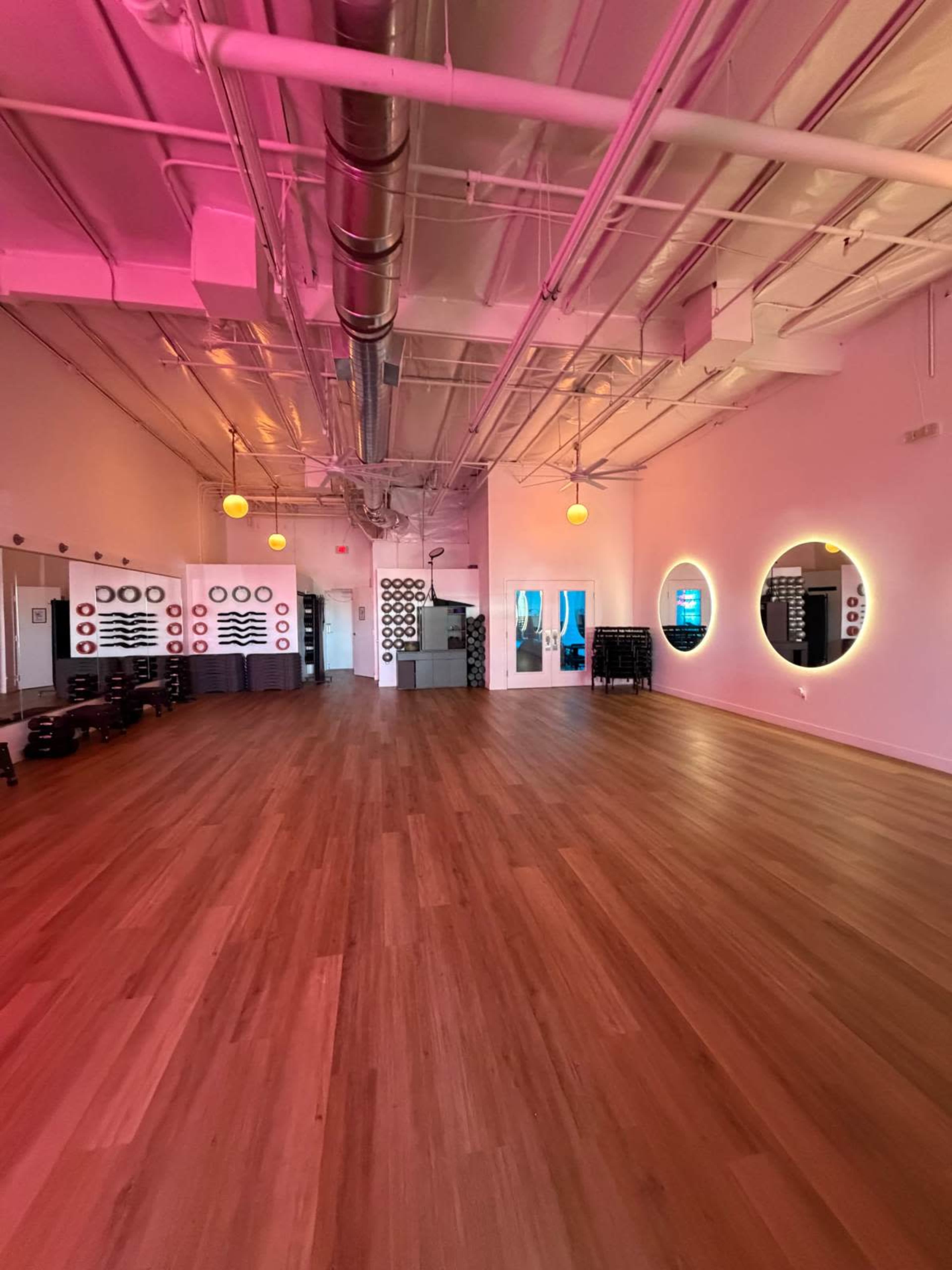The image shows a spacious, modern studio with wooden flooring, pink lighting, and circular mirrors on the walls.
