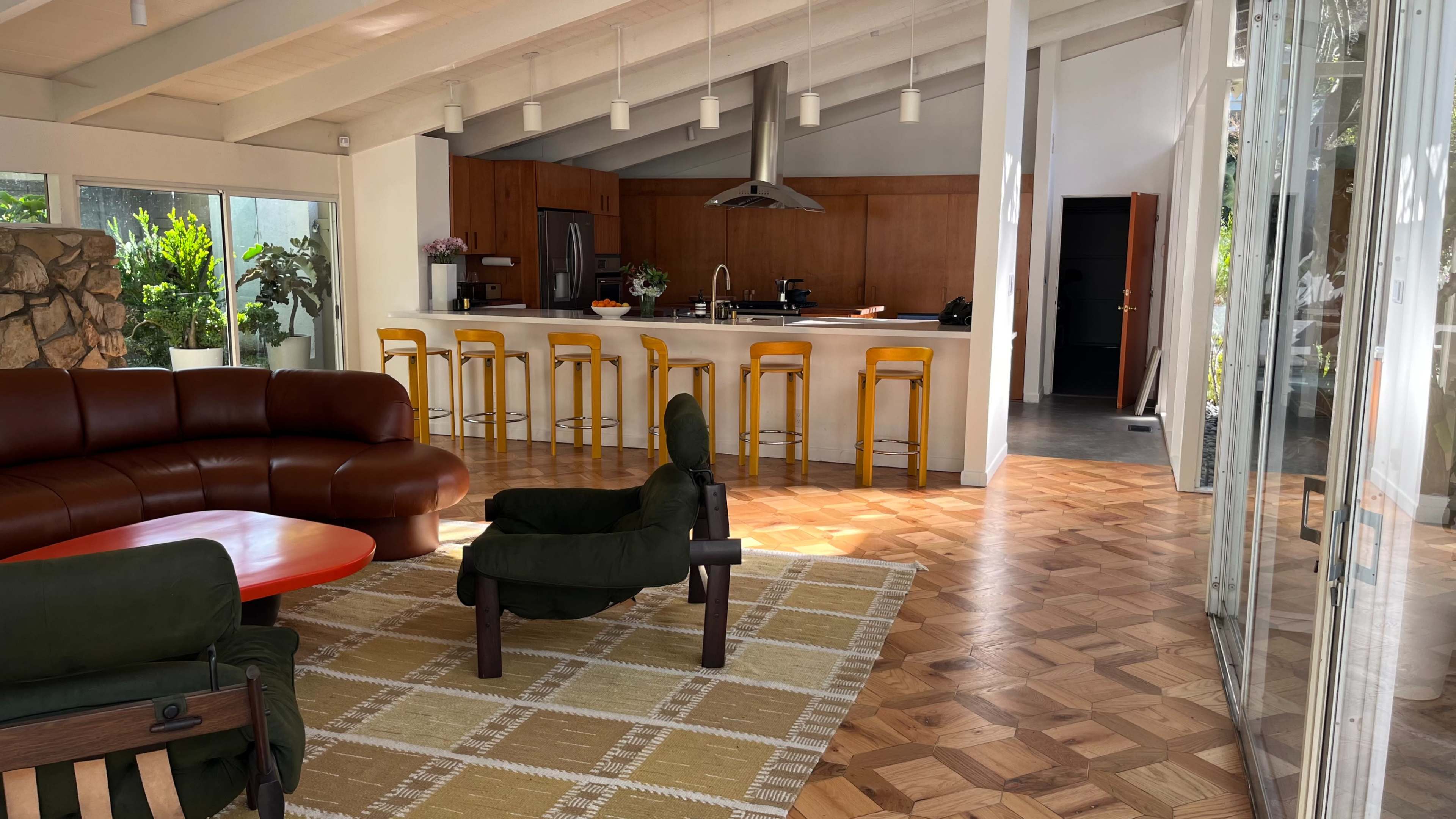 The image shows a modern, open-concept kitchen and living area featuring a brown leather sofa, a green armchair, and a large bar with yellow stools.