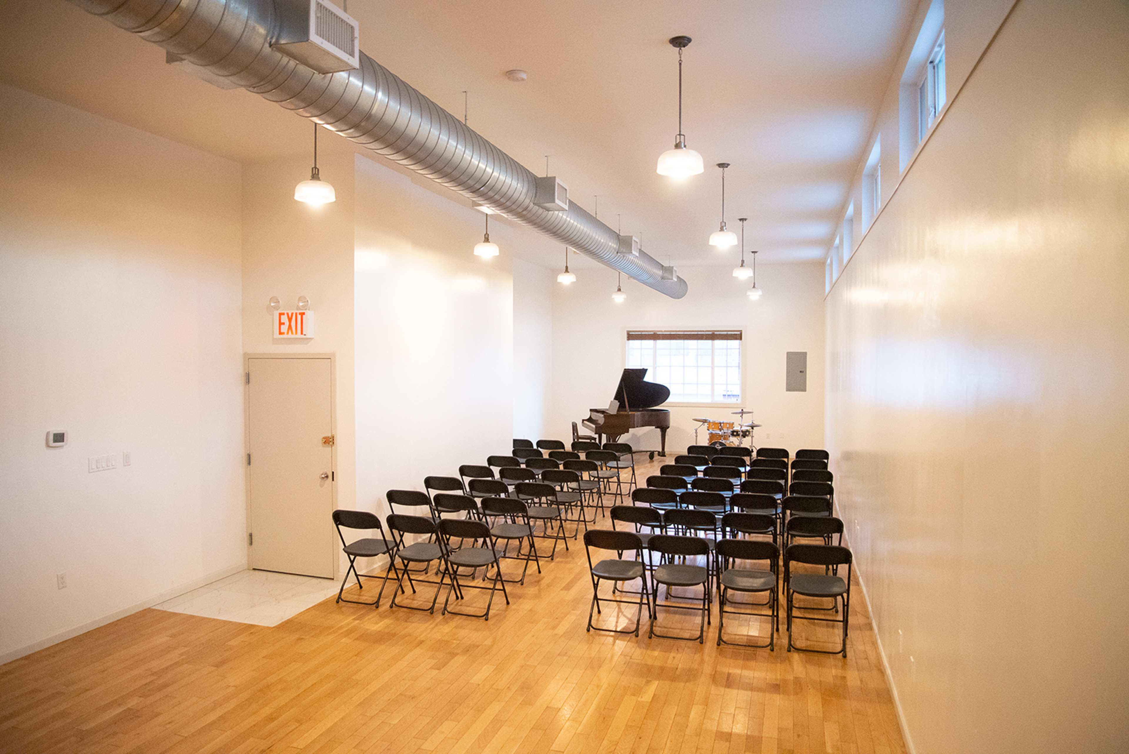 A small performance space features a grand piano and rows of black folding chairs arranged on a wooden floor.