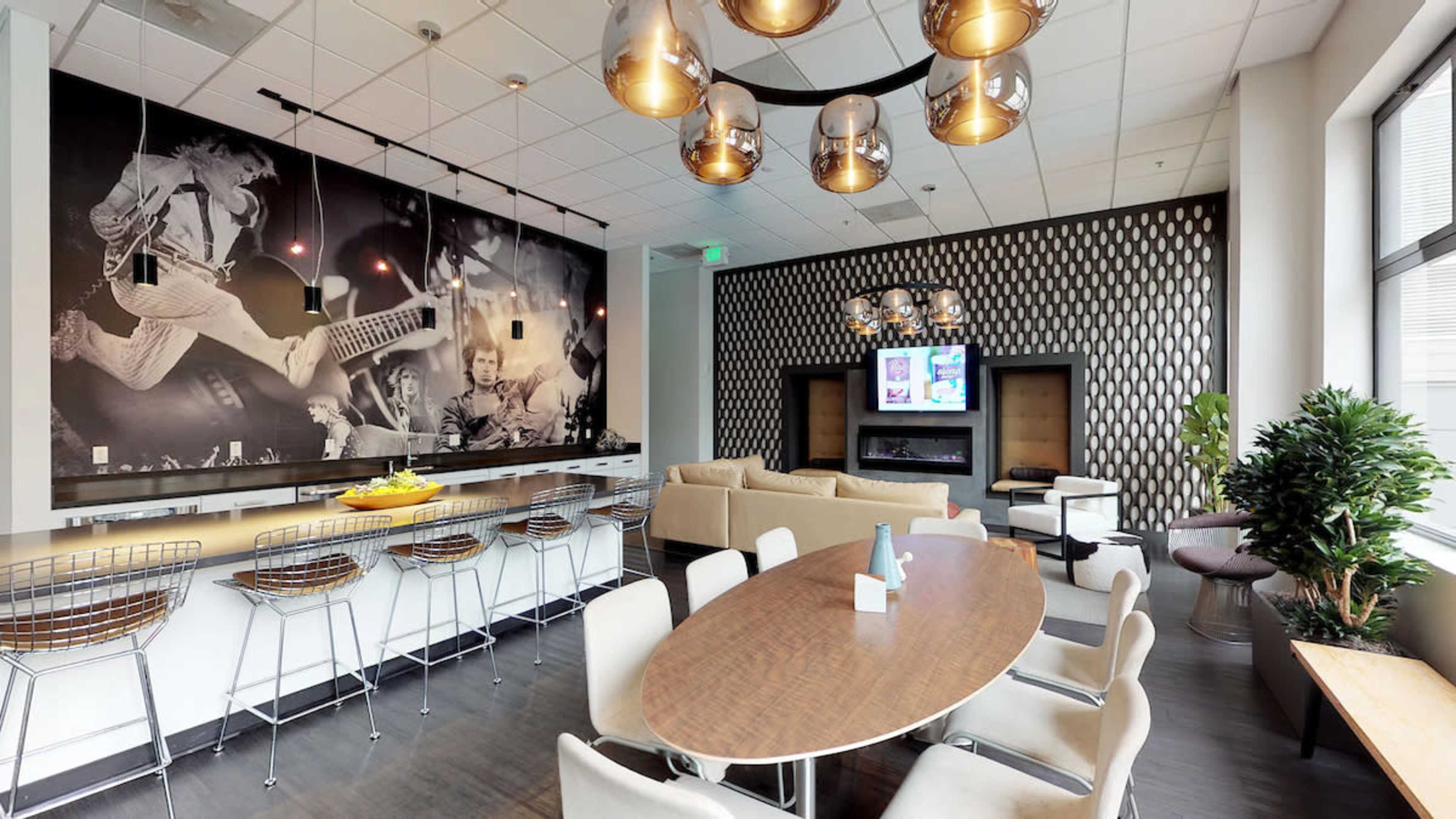 A modern lounge area featuring a large wall mural of musicians, a long oval dining table, bar seating, and stylish lighting fixtures.
