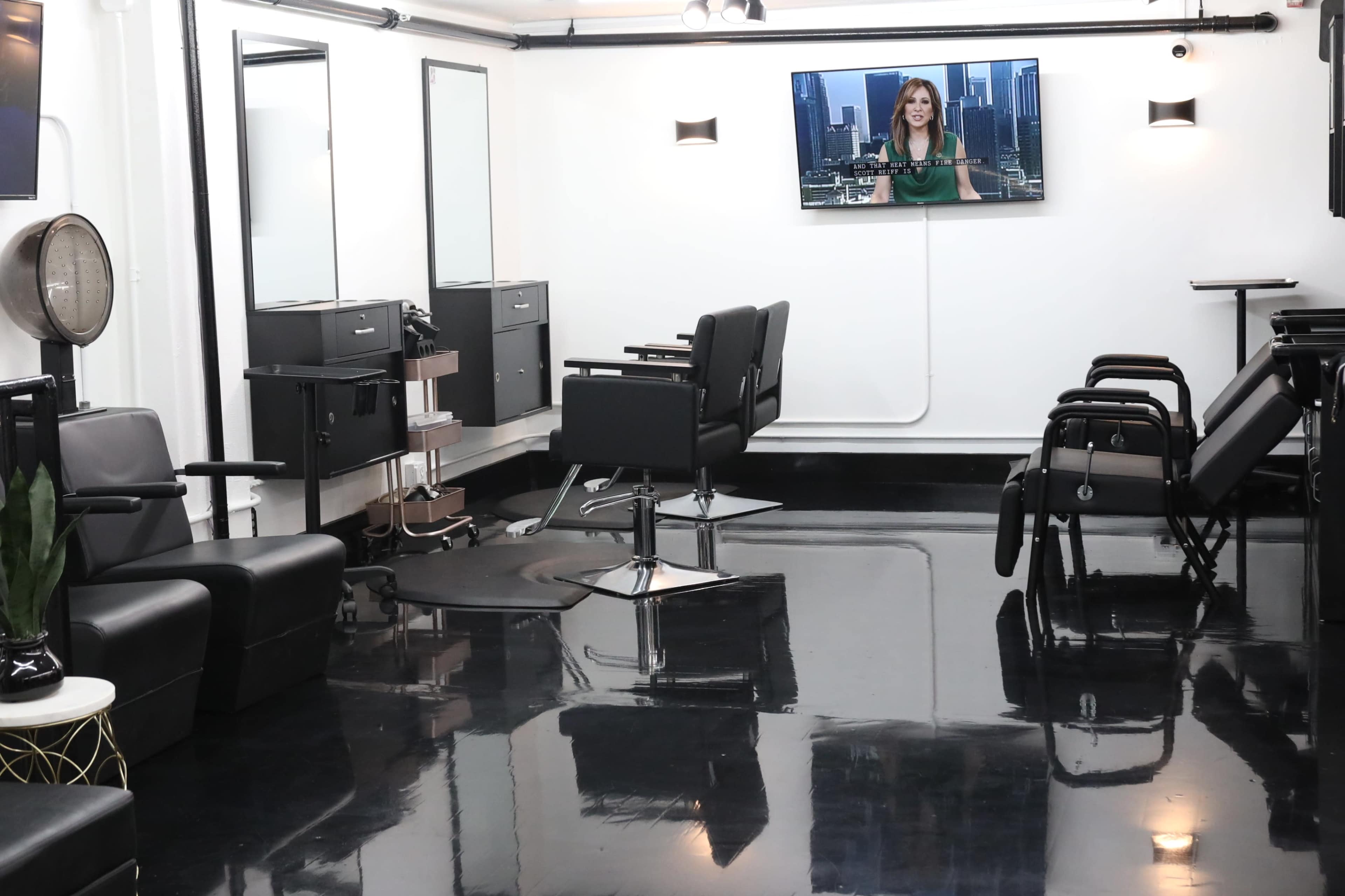 A modern hair salon interior with black chairs, mirrors, and a television mounted on the wall displaying a news broadcast.