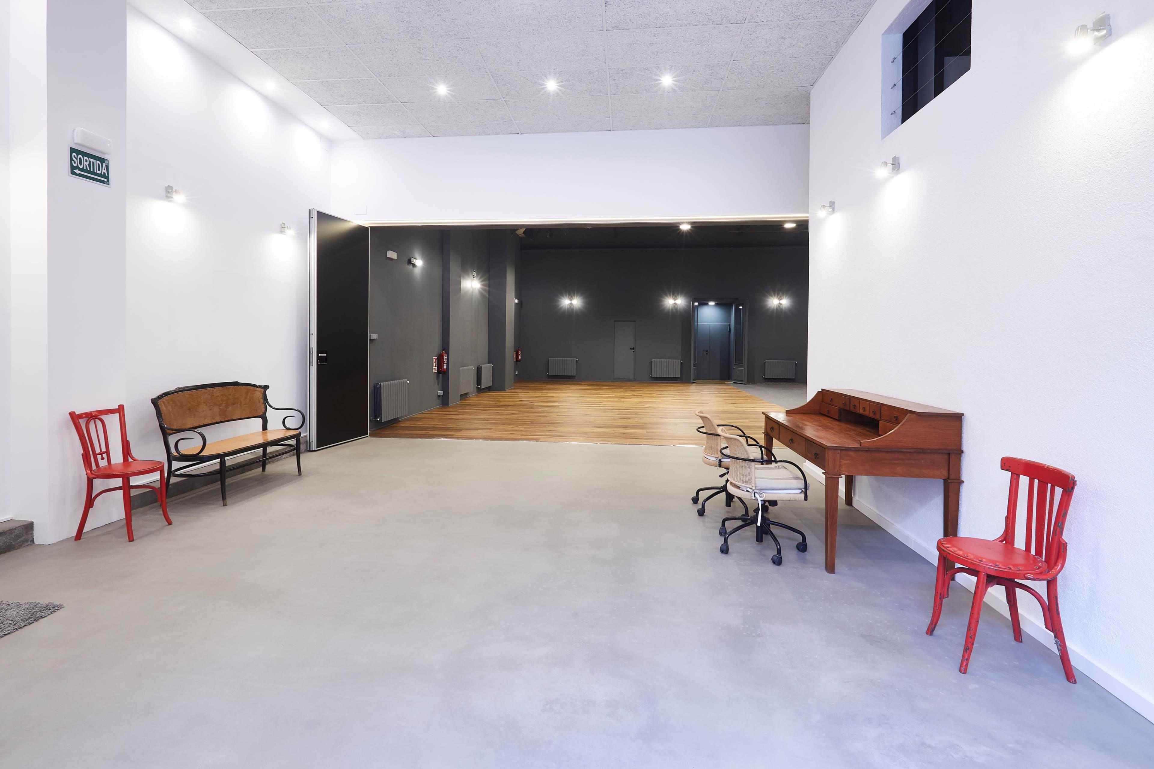 An empty indoor space featuring wooden flooring, white walls, and a simple seating arrangement with a wooden desk and chairs in contrasting colors.