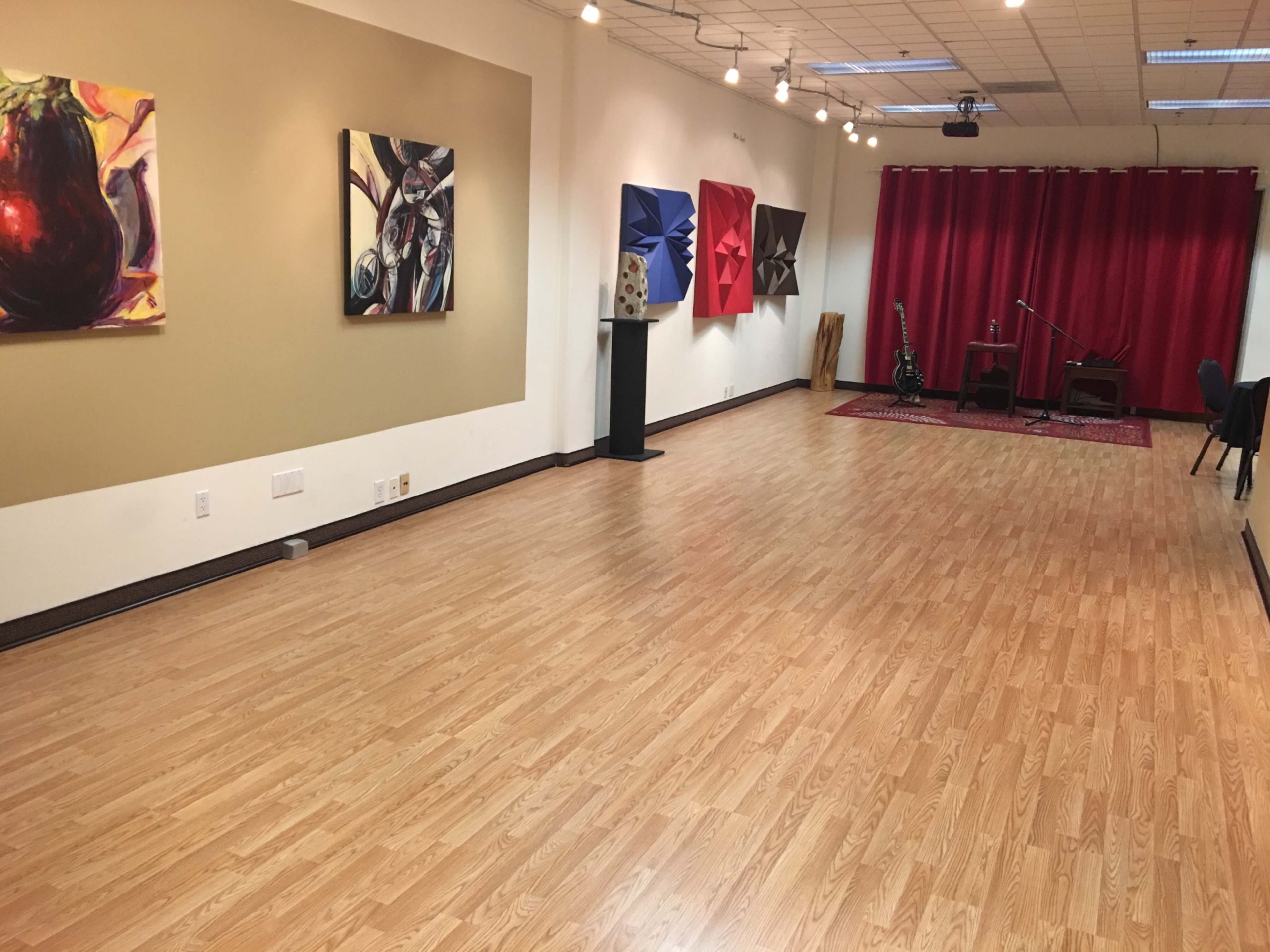 The image shows a spacious room with wooden flooring, featuring colorful artwork on the walls and a small stage area with a microphone and musical instruments.