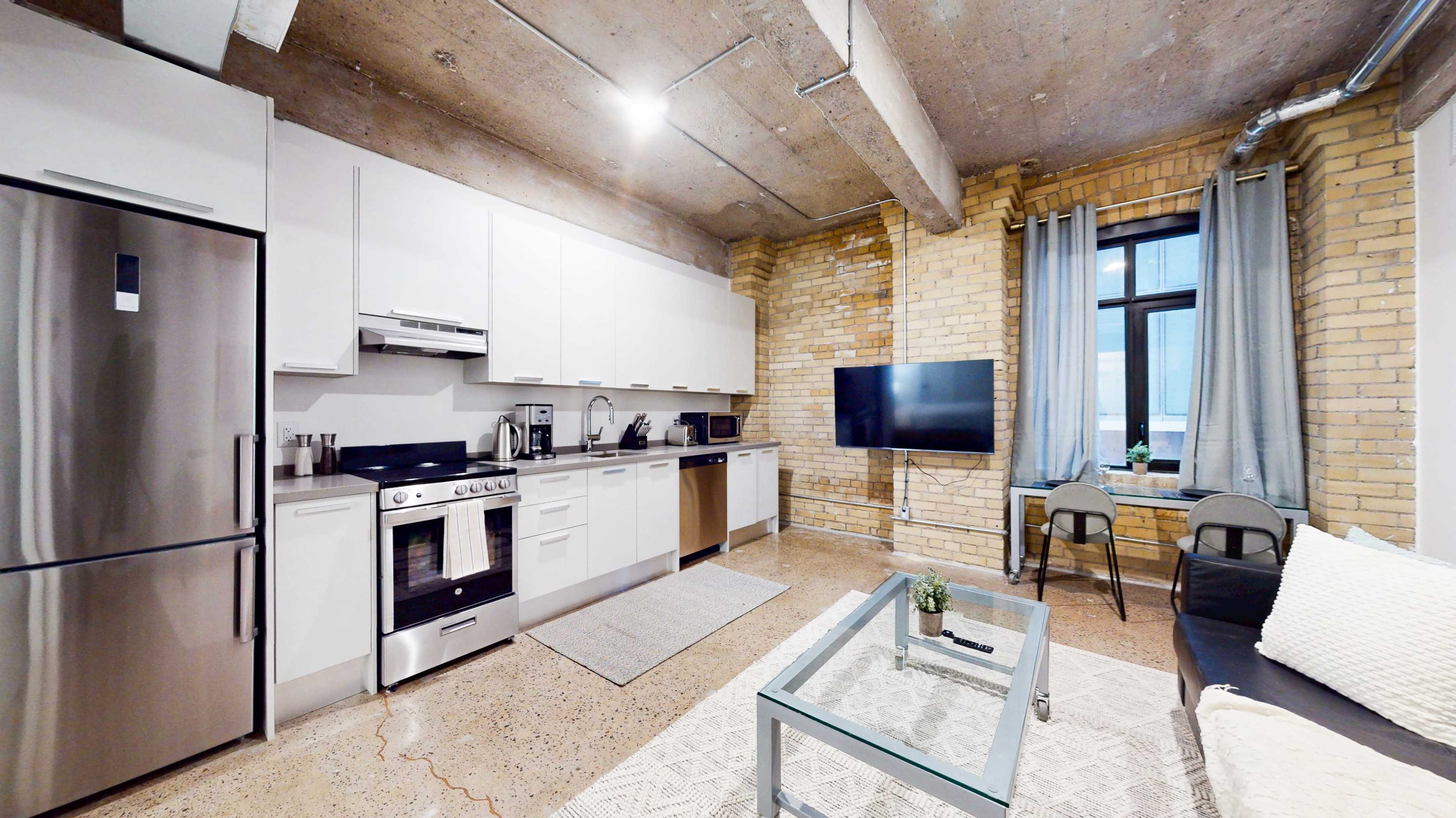The image shows a modern kitchen and living area with stainless steel appliances, a brick wall, and a large window.