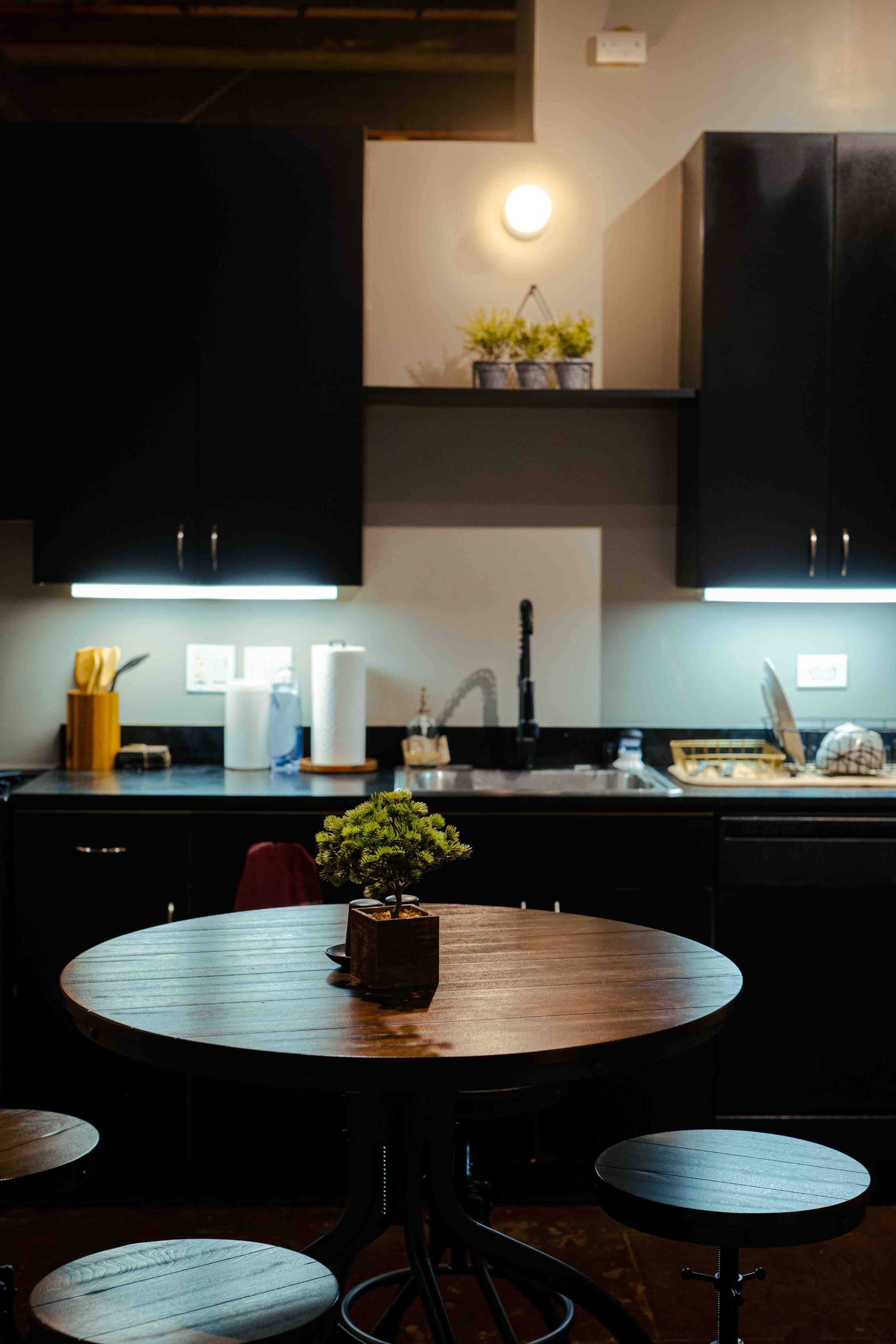 A round wooden table with stools is positioned in a kitchen featuring black cabinets and a sink under wall-mounted lights.