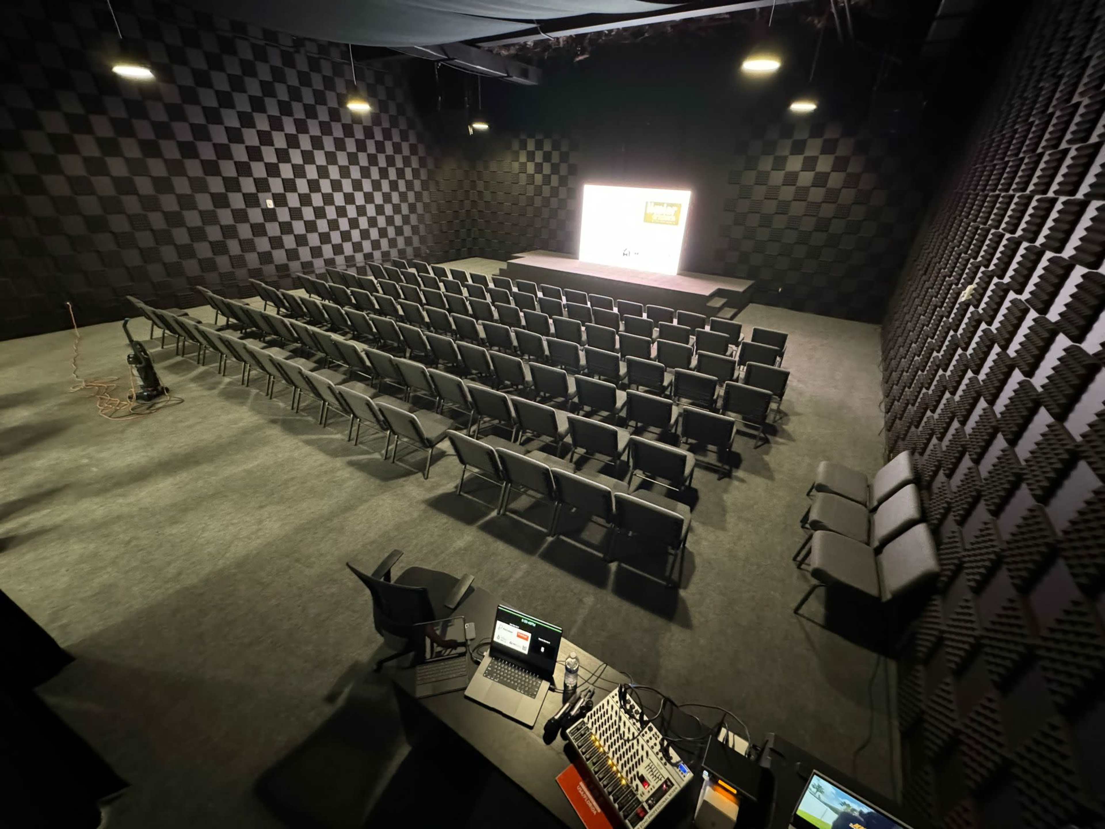 The image shows a spacious auditorium with rows of empty chairs facing a projector screen, set against a textured black wall.