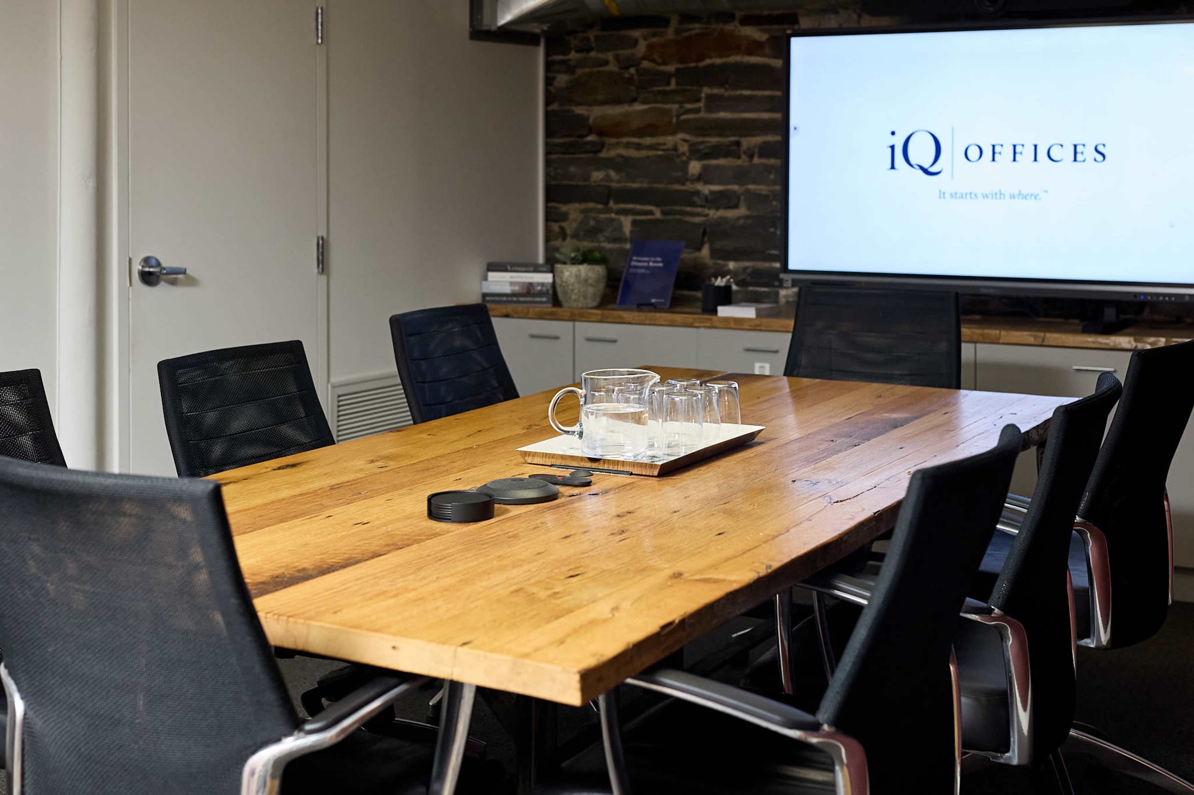 A modern conference room features a large wooden table surrounded by black chairs, with a television screen displaying the iQ Offices logo.