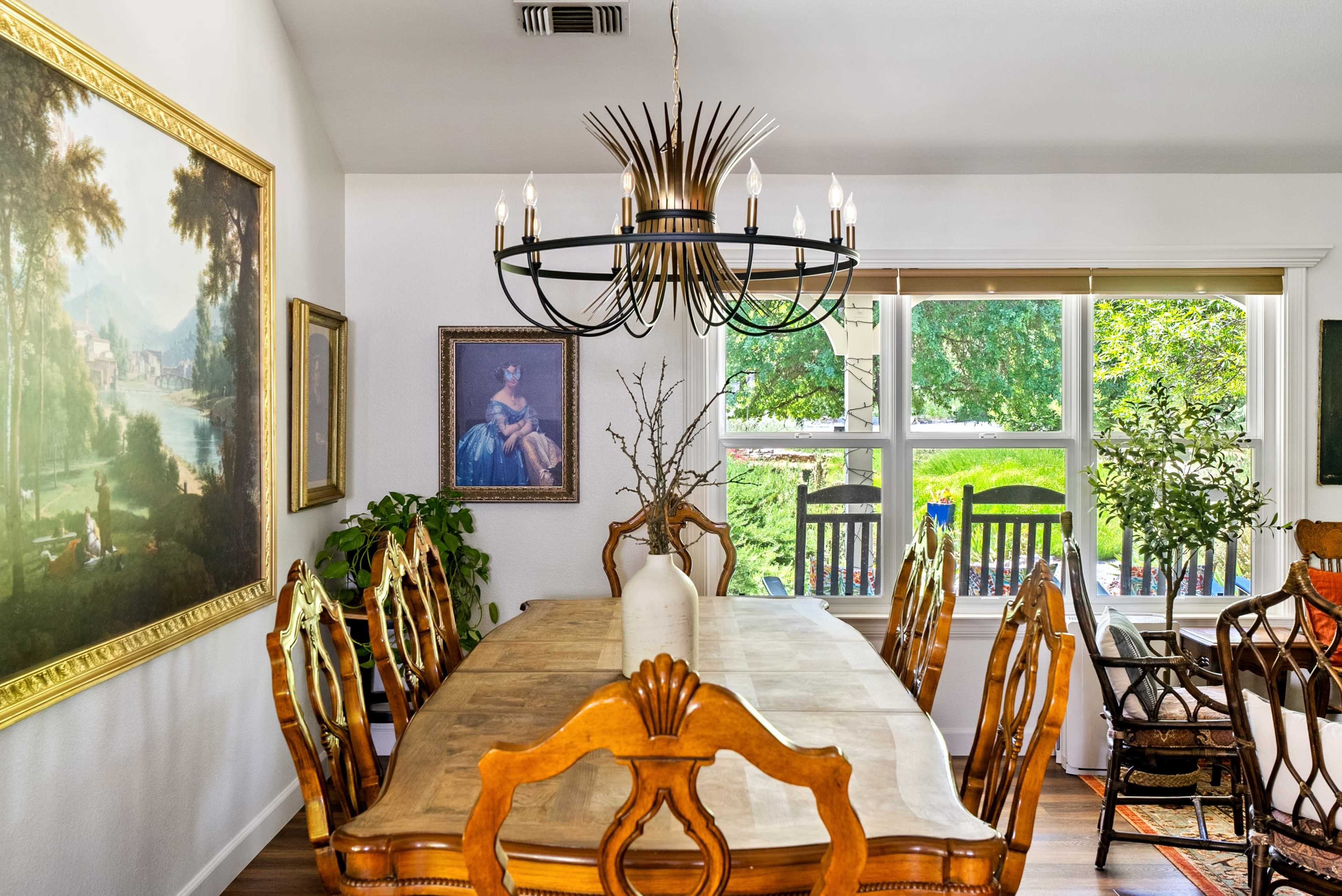 A wooden dining table with ornate chairs is set under a large chandelier in a well-lit room featuring decorative artwork and a view of greenery outside.