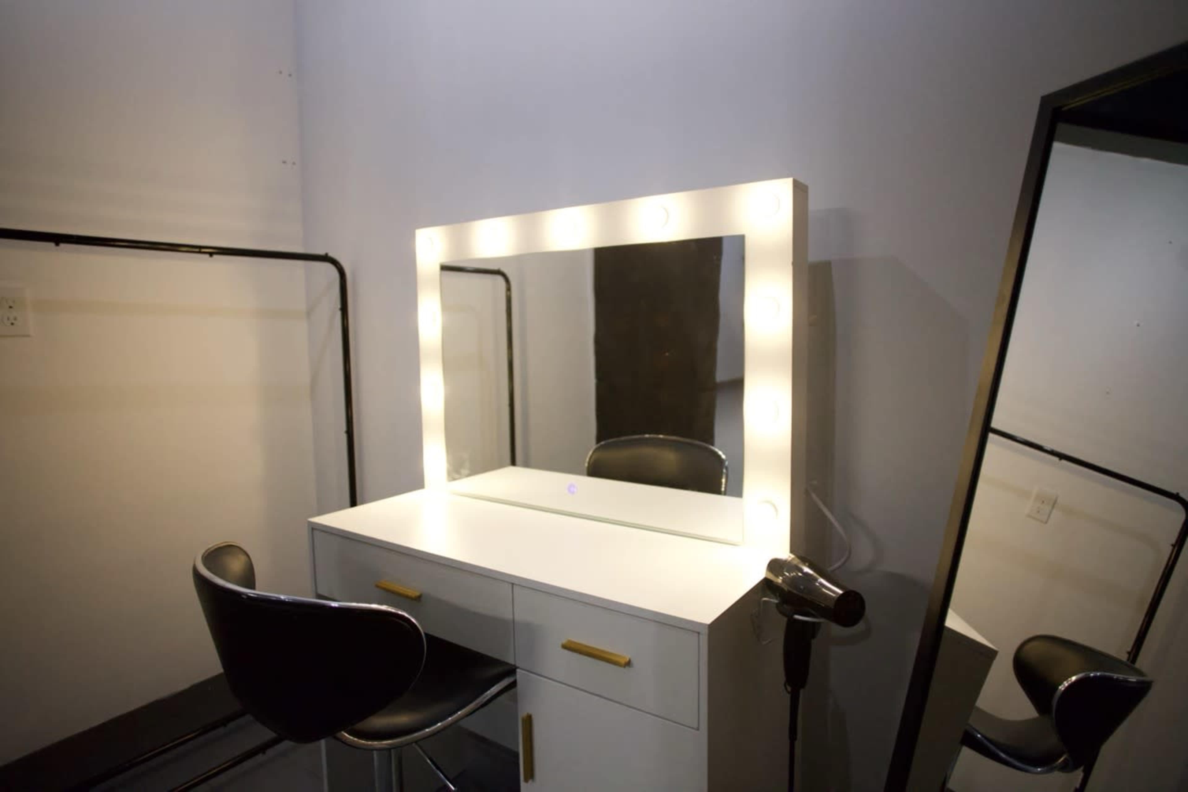 The image shows a modern makeup vanity with a large illuminated mirror, a black stool, and a hairdryer next to it, set against a plain wall.