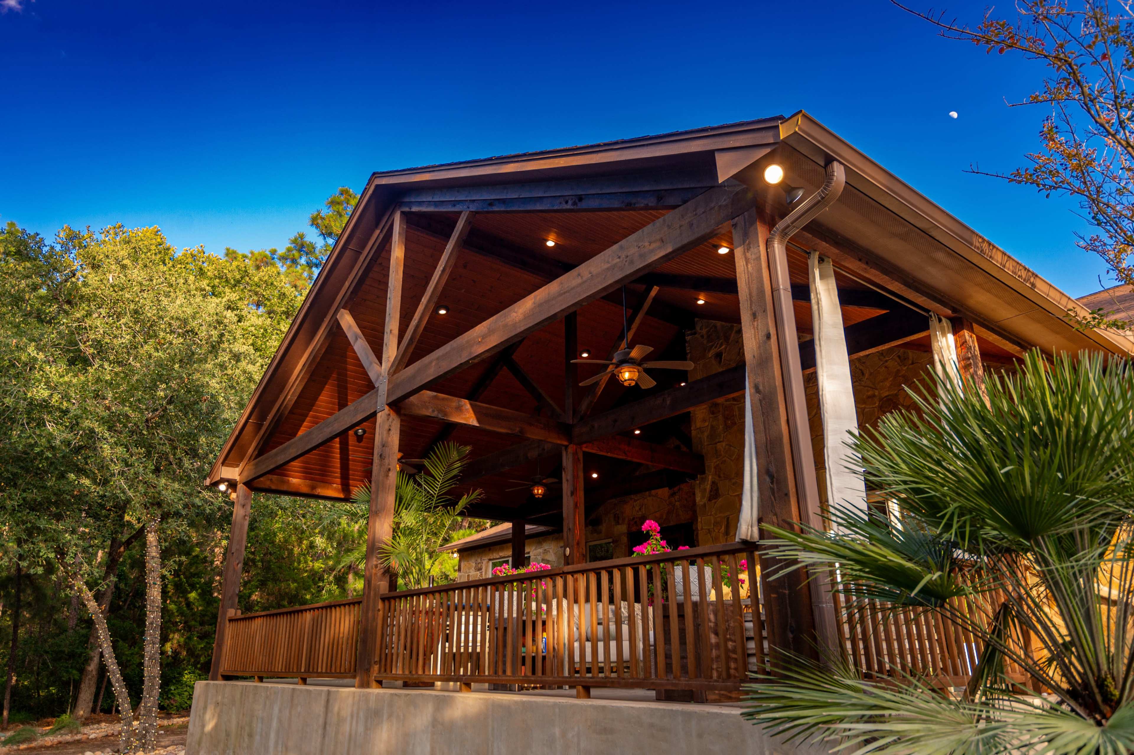 The image shows a wooden porch with a gabled roof supported by beams, surrounded by greenery and featuring potted plants.