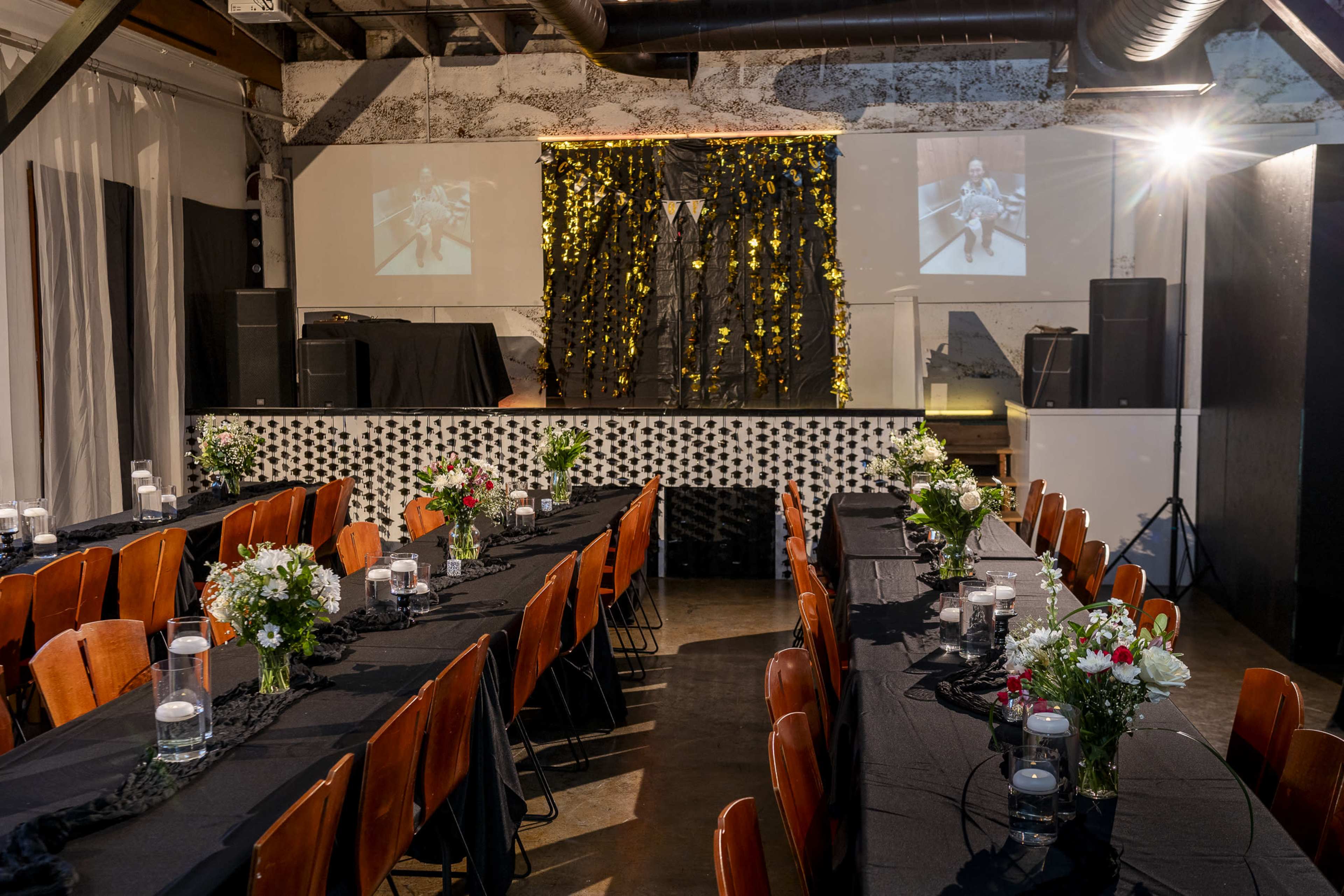 The image shows a banquet hall set for an event, featuring long tables adorned with black tablecloths and centerpieces of flowers, with a stage in the background decorated with curtains and screens.