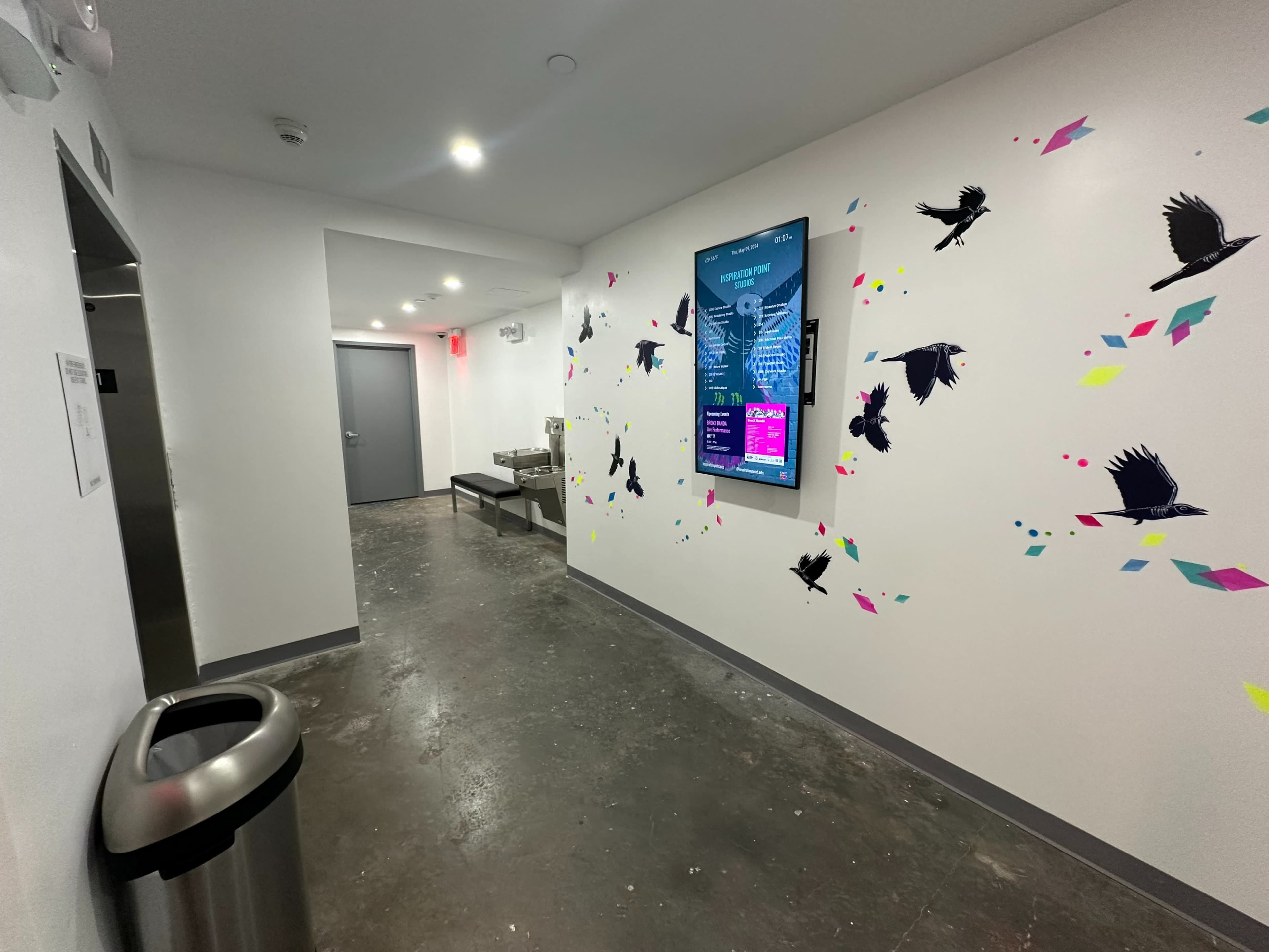 The image shows a hallway with a wall displaying colorful bird decals, a digital screen mounted above, and a trash can at the corner.