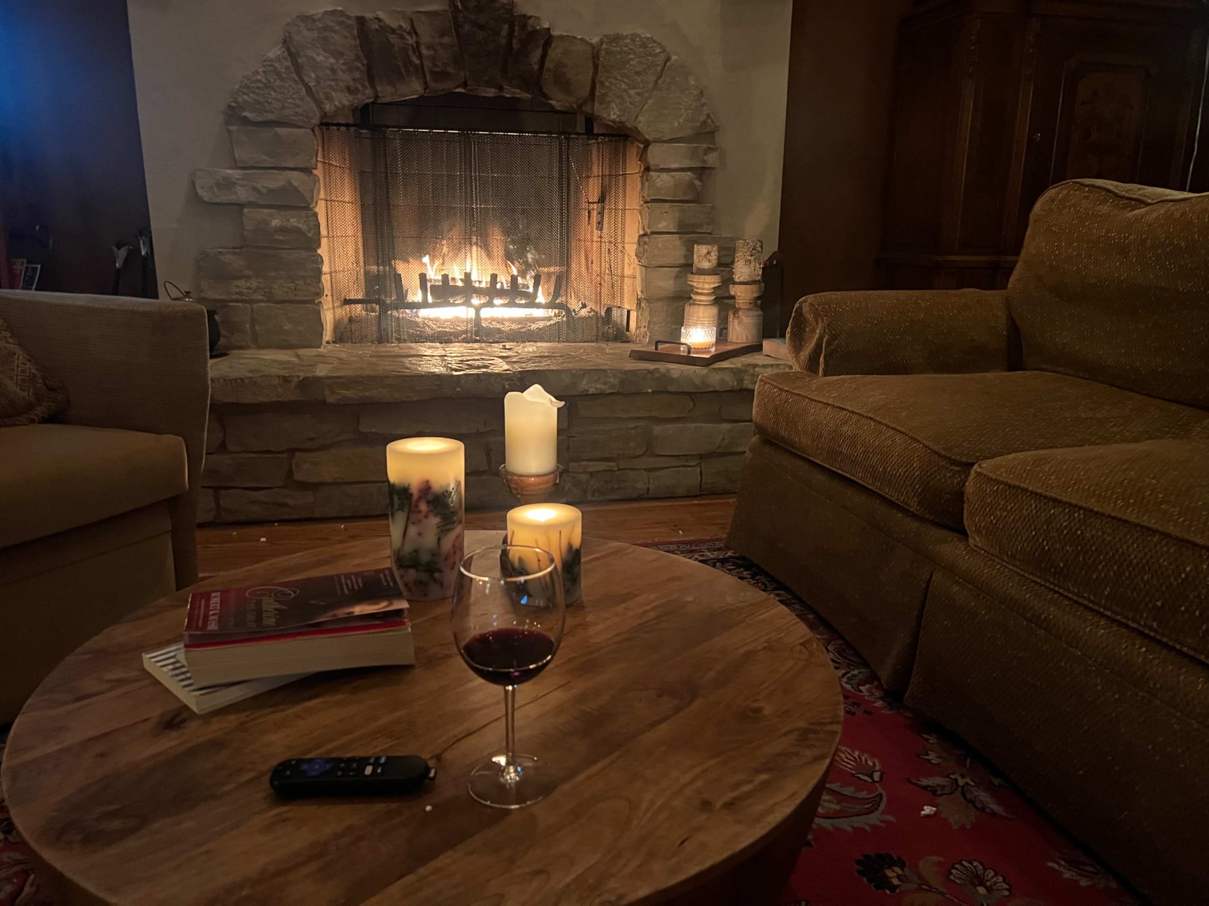 A cozy living room features a stone fireplace with a glowing fire, a wooden coffee table with a book, a remote, and a glass of red wine, surrounded by candles.