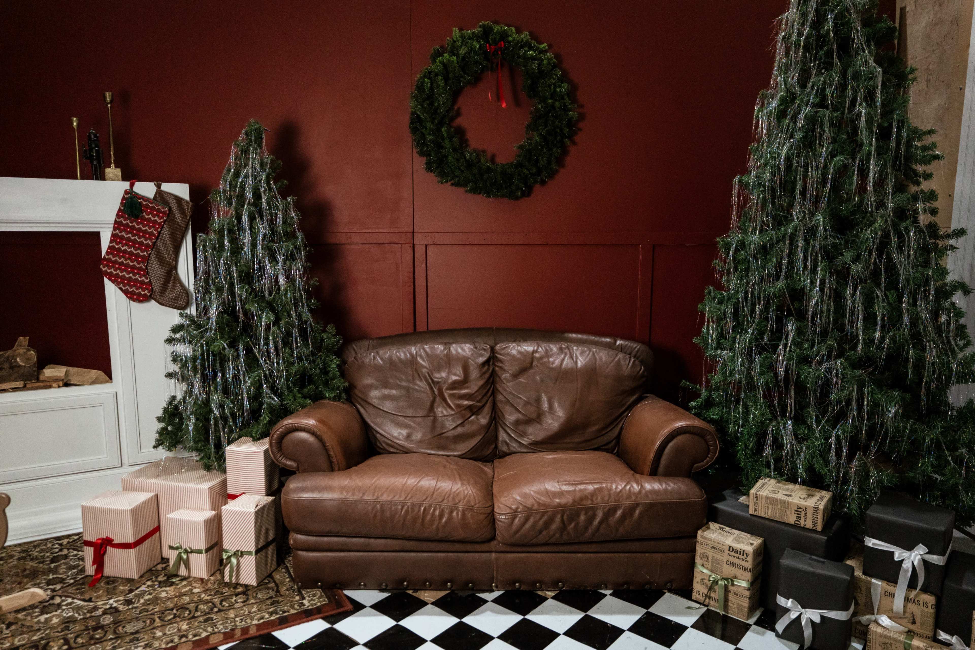 A brown leather couch is positioned in front of a red wall, flanked by decorated Christmas trees and surrounded by wrapped gifts.