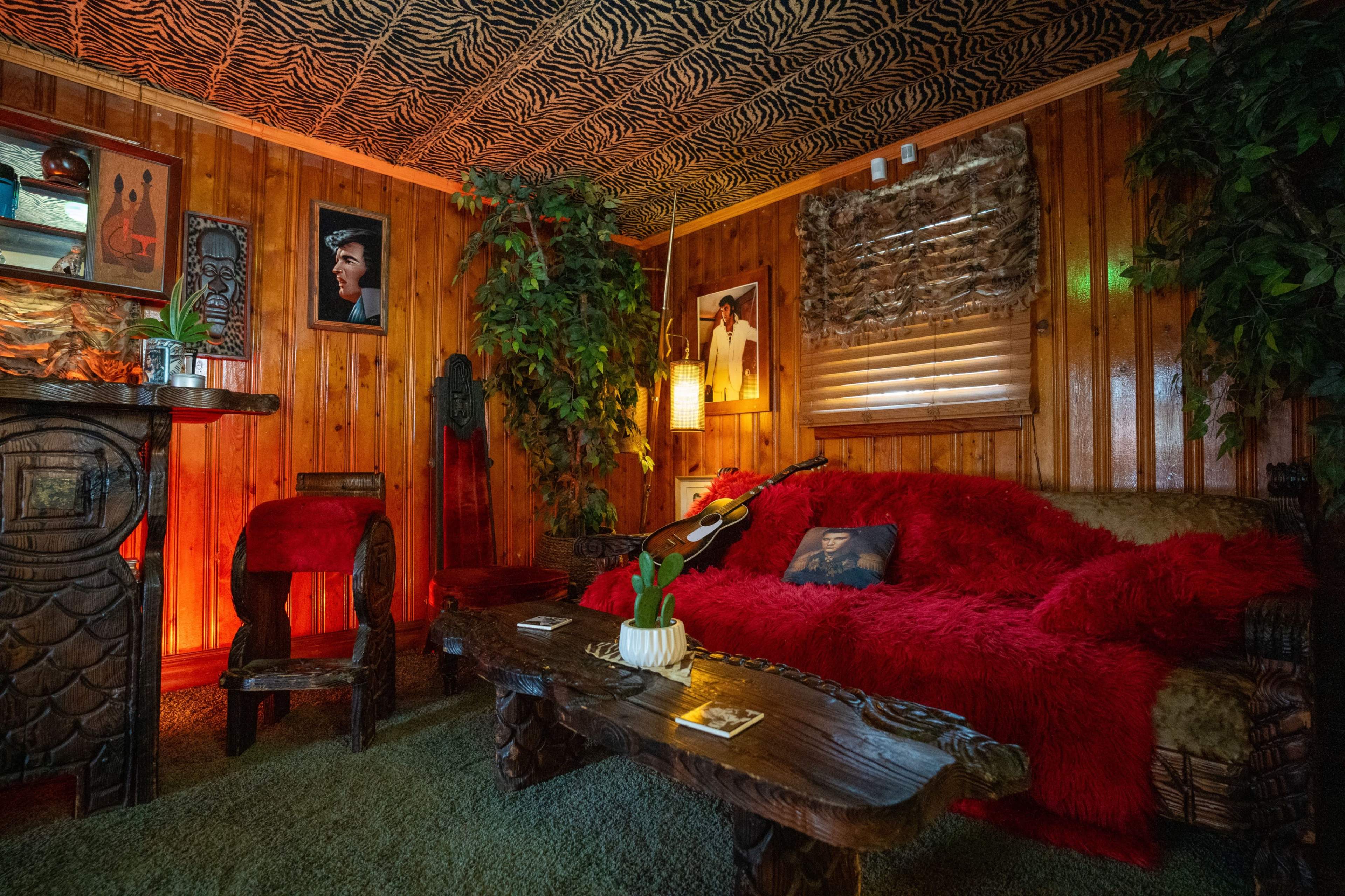 A warmly lit, rustic room with wooden walls, a textured ceiling, a red throw blanket on a couch, and decorative plants alongside artwork on the walls.