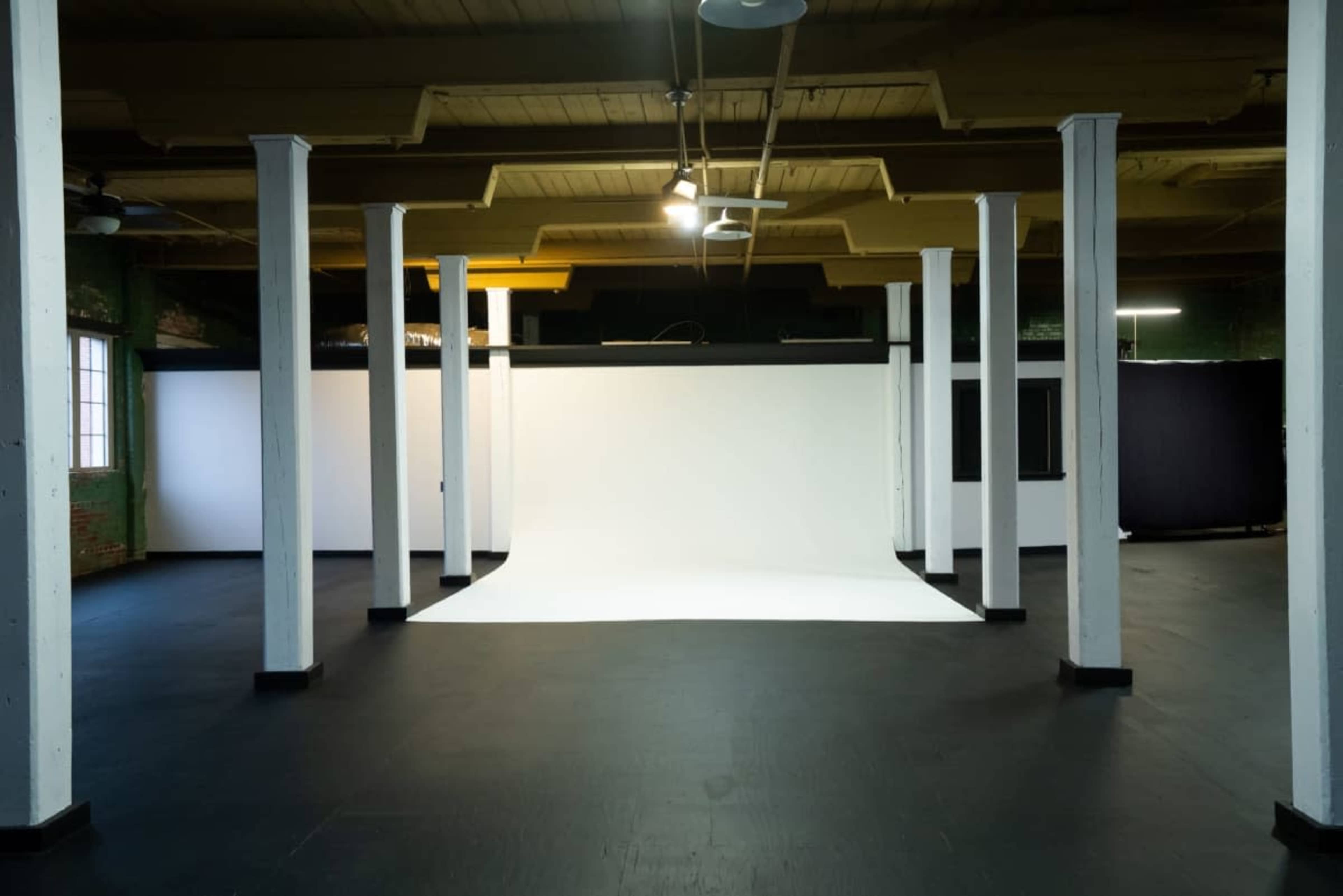 The image shows an empty photography studio with a white backdrop, black curtains, and exposed columns under dim lighting.