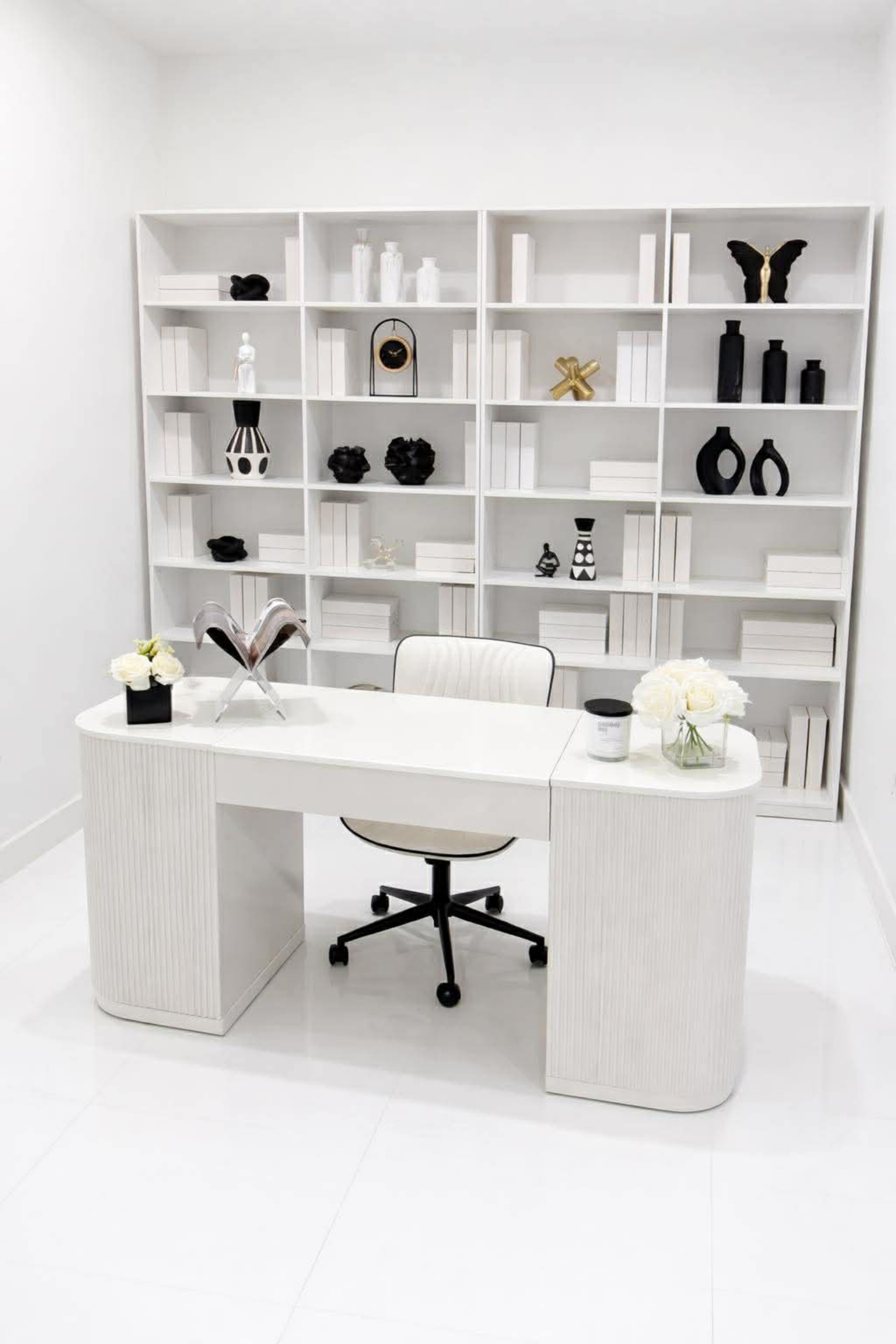The image shows a modern white office space with a sleek desk and an organized bookshelf filled with decorative objects.